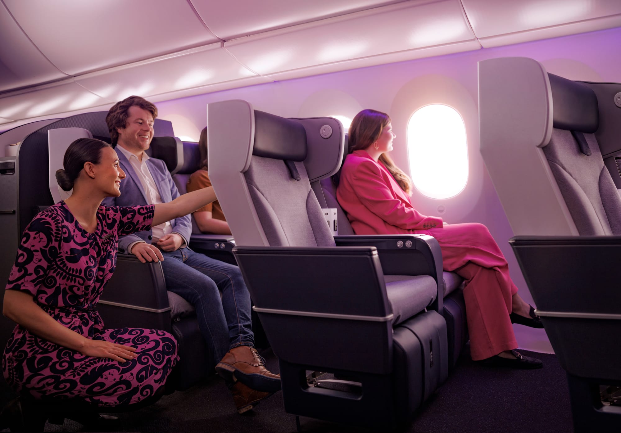 Interior, airline cabin, premium economy seats. People sit in seats and a flight attendant is pointing something out to a passenger. At the centre is a premium economy seat with legrest and a fixed back.