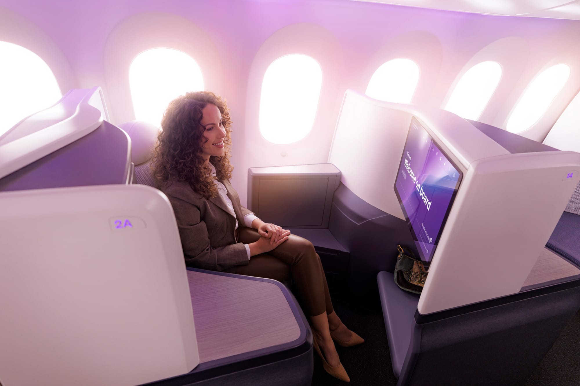 Interior, airline cabin. A woman sits in the inward-facing herringbone seat at the window.