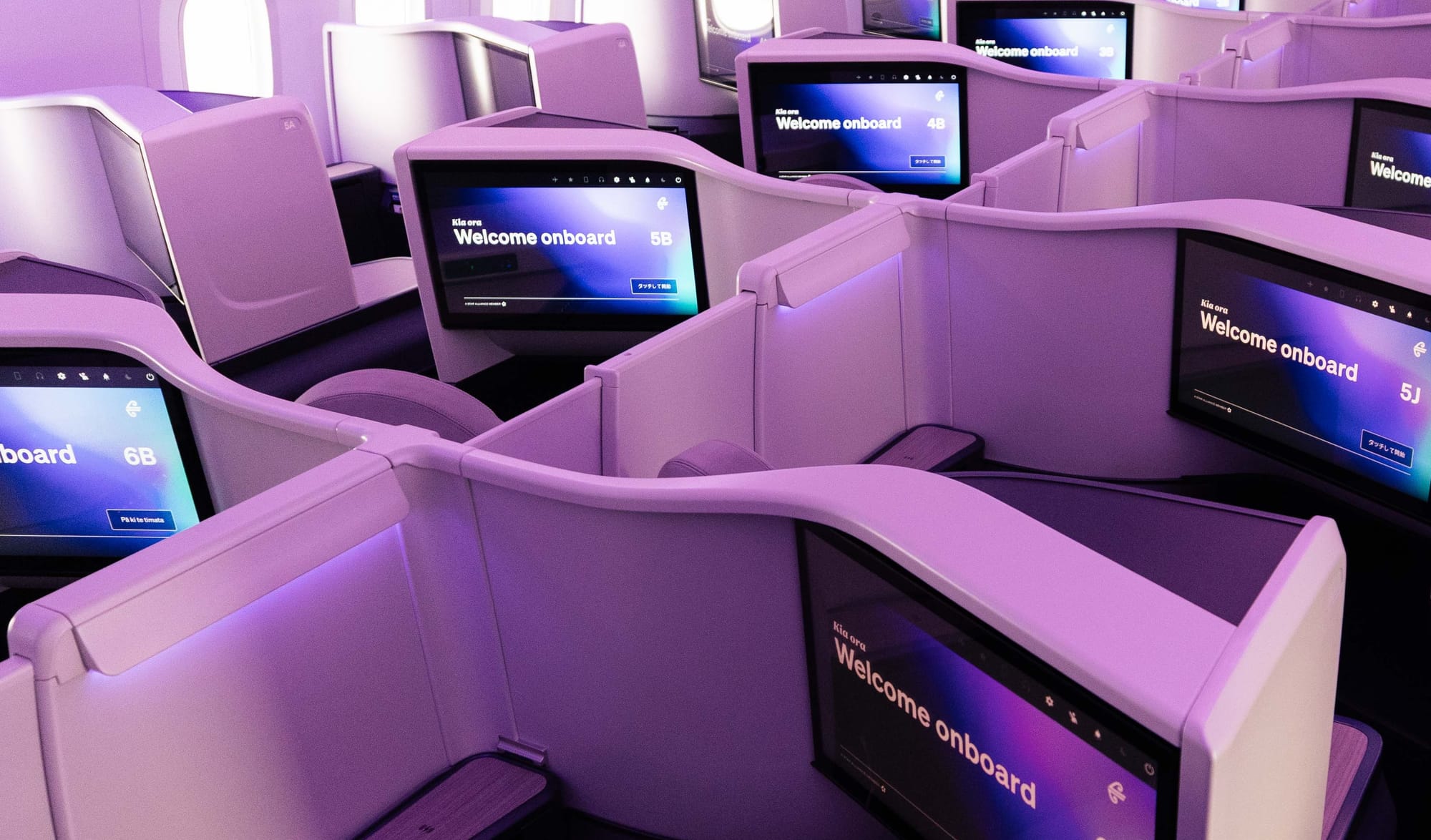Closeup, airline cabin, showing the centre pairs and some of the outboard seats. There's a lot of purple-washed off-white thermoplastic at varying different angles and shapes.