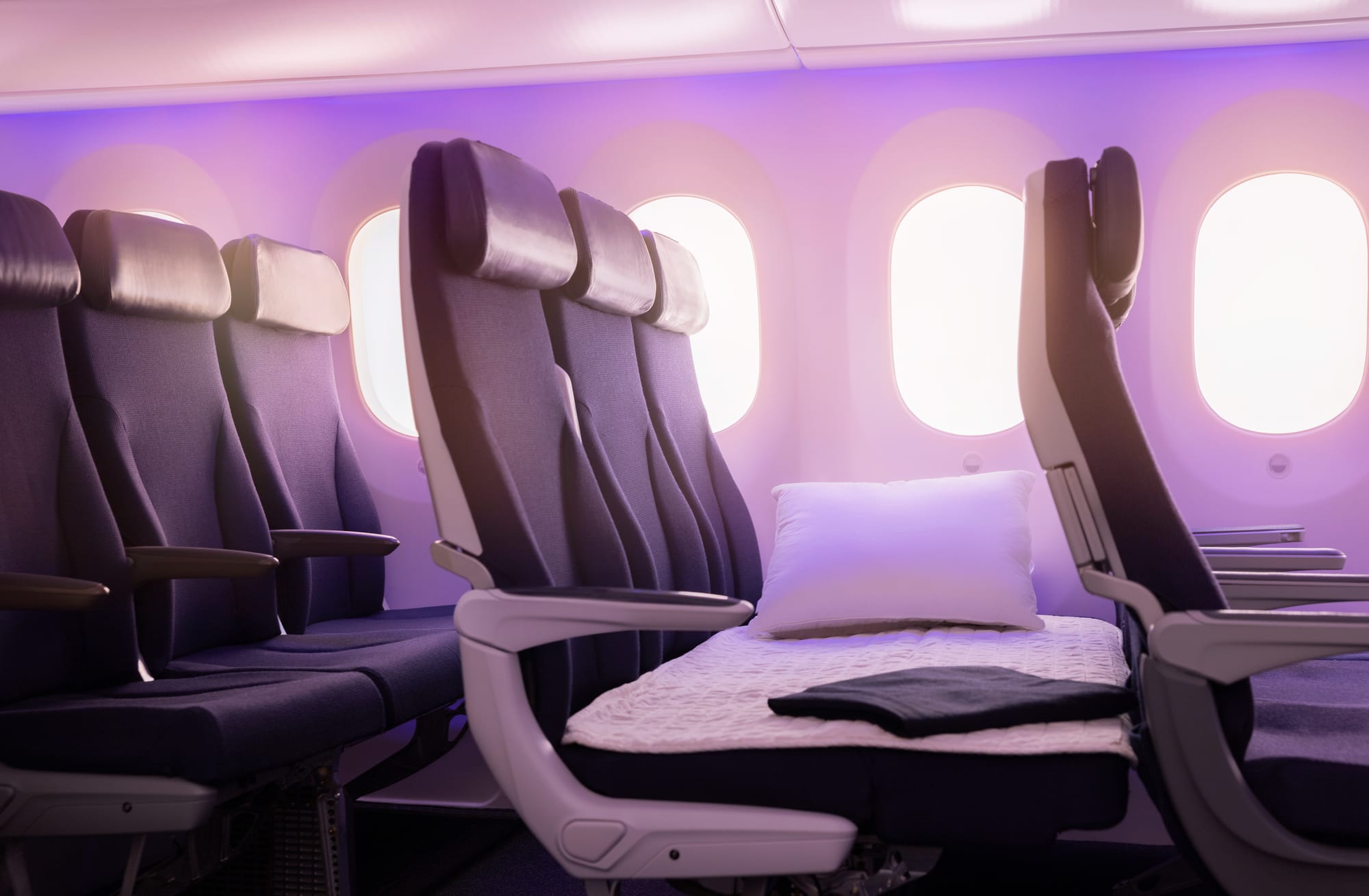 Interior, airline cabin, economy row of 3 by a window. The legrests of the seats have extended upwards to create a flat surface, like a very short bed, which is covered with a mattress pad, and there is also a pillow.