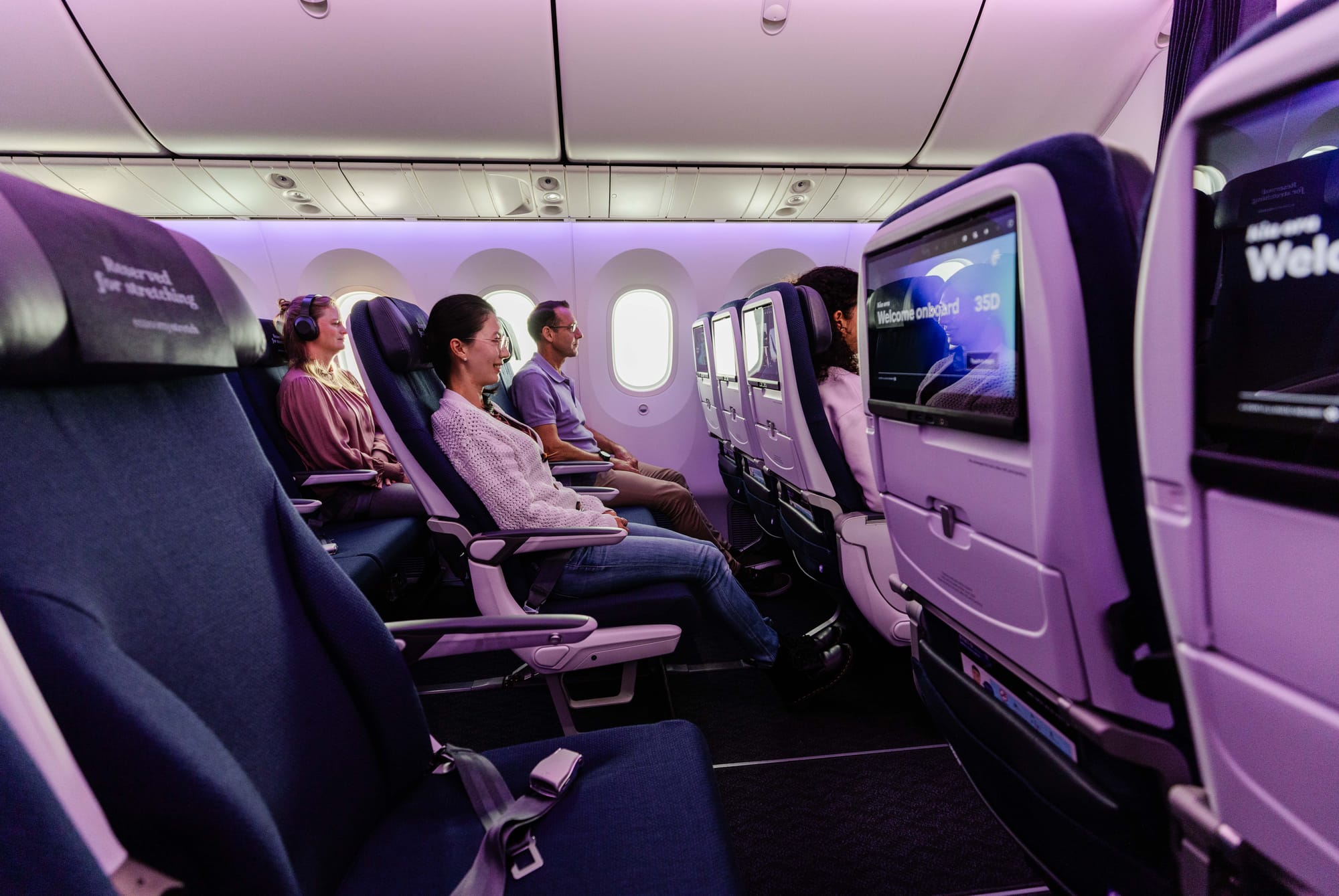 Interior, airline cabin, economy. A very short woman is seated in the focus seat, a C-position aisle seat on the window side of the aisle. Her head is below the top of the headrest, and her knees barely extend to the end of the seat cushion.