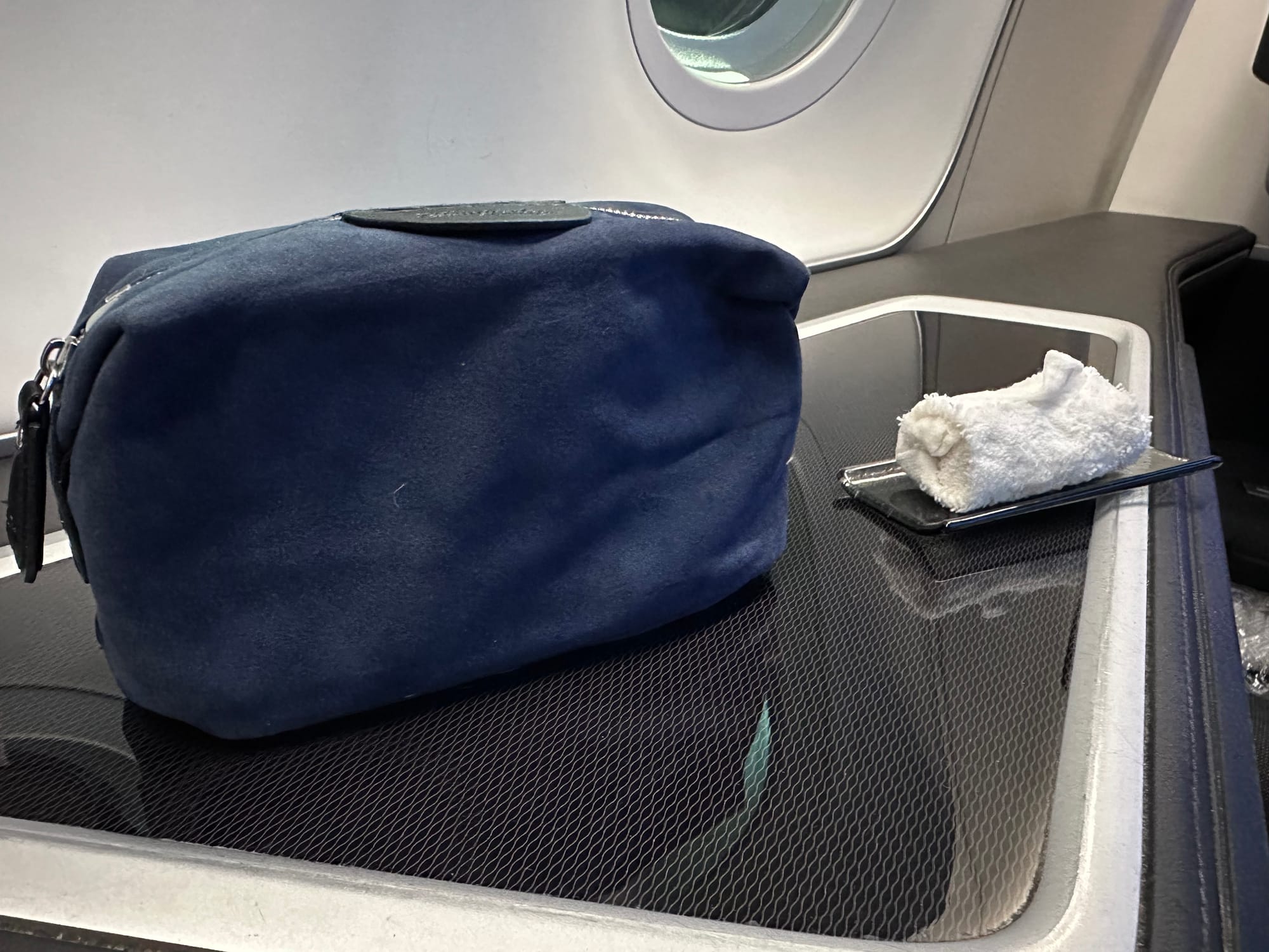 Closeup, first class seat side table. A squashed-looking soft sided dark blue suede-effect amenity kit sits next to a tiny white hand towel that is rolled very tightly and scrunched up.