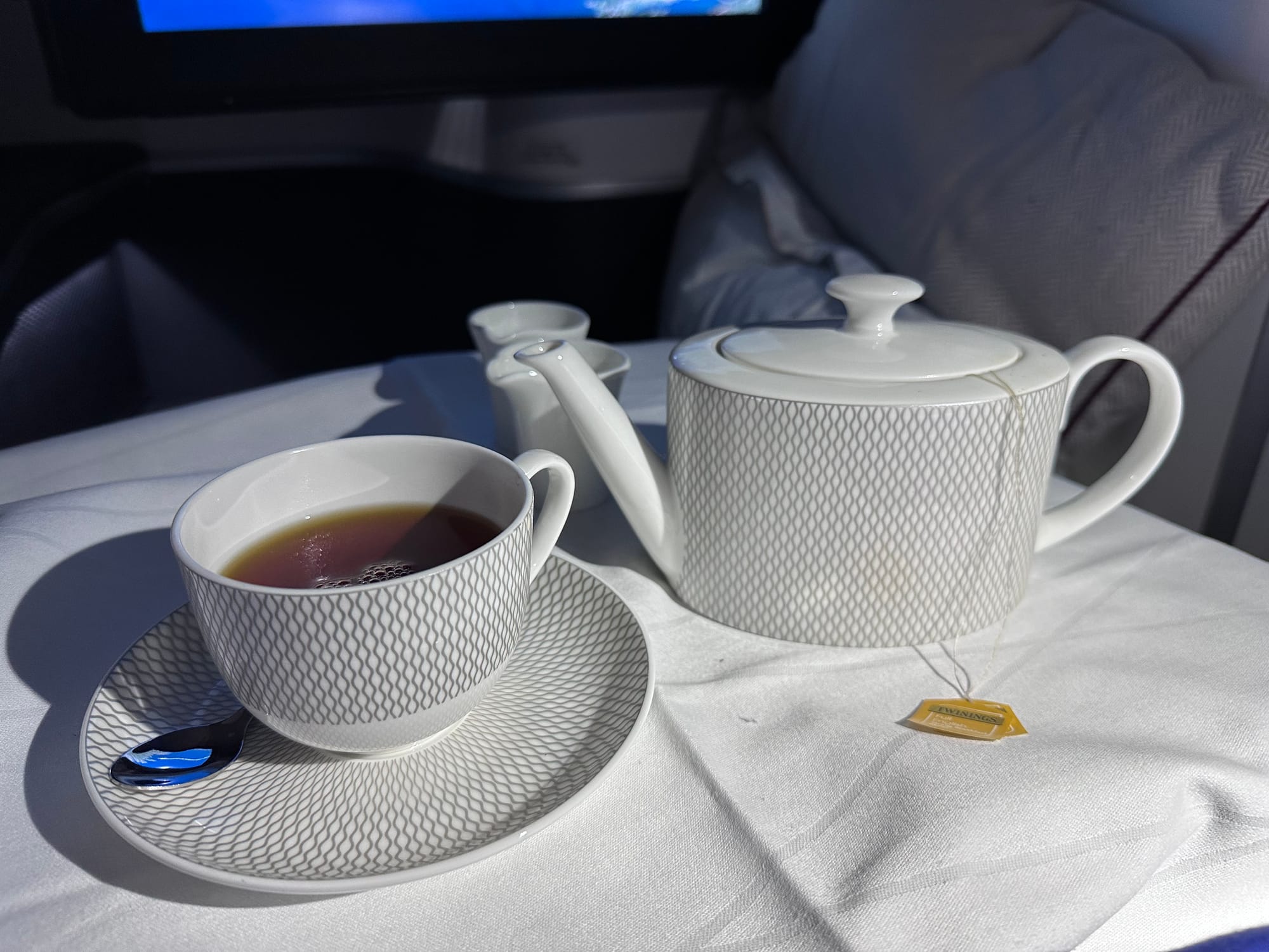 Close up, first class tray table with white polyester-cotton blend tablecloth. A rather thick teacup full of black tea, on a saucer, both with a ribbed pattern sit next to a teapot in the same pattern that has Twinings tea brand tags coming out of it. Behind are two tiny milk pots.