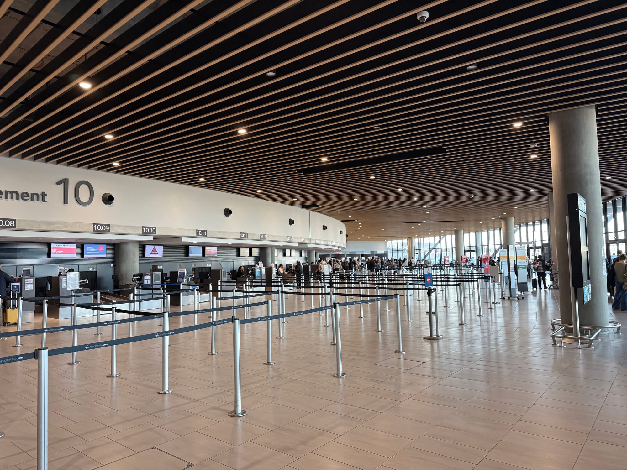 Interior, Lyon Airport departures area. Wooden slatted roof in a circular well-lit atrium. Many tensa-barrier queues are in place with several small signs for various airlines. They are not legible, but especially illegible is the Lufthansa placard, which is white on yellow.