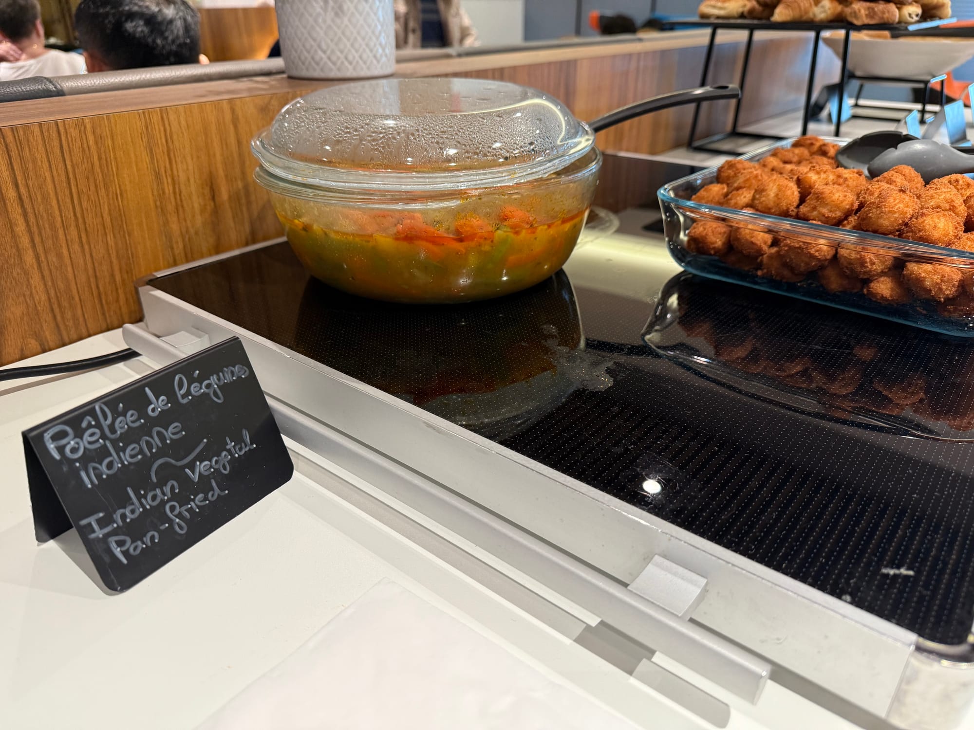 Closeup, lounge food. It is incredibly unappealing: frozen-and-unfrozen-looking vegetables swimming in a vomit-green stew with a film of oil on top, and a large tray of inch-diameter fried balls that are cod. A handwritten sign notes: "Poêleé de légumes indienne ~ Indian vegetal Pan-fried".