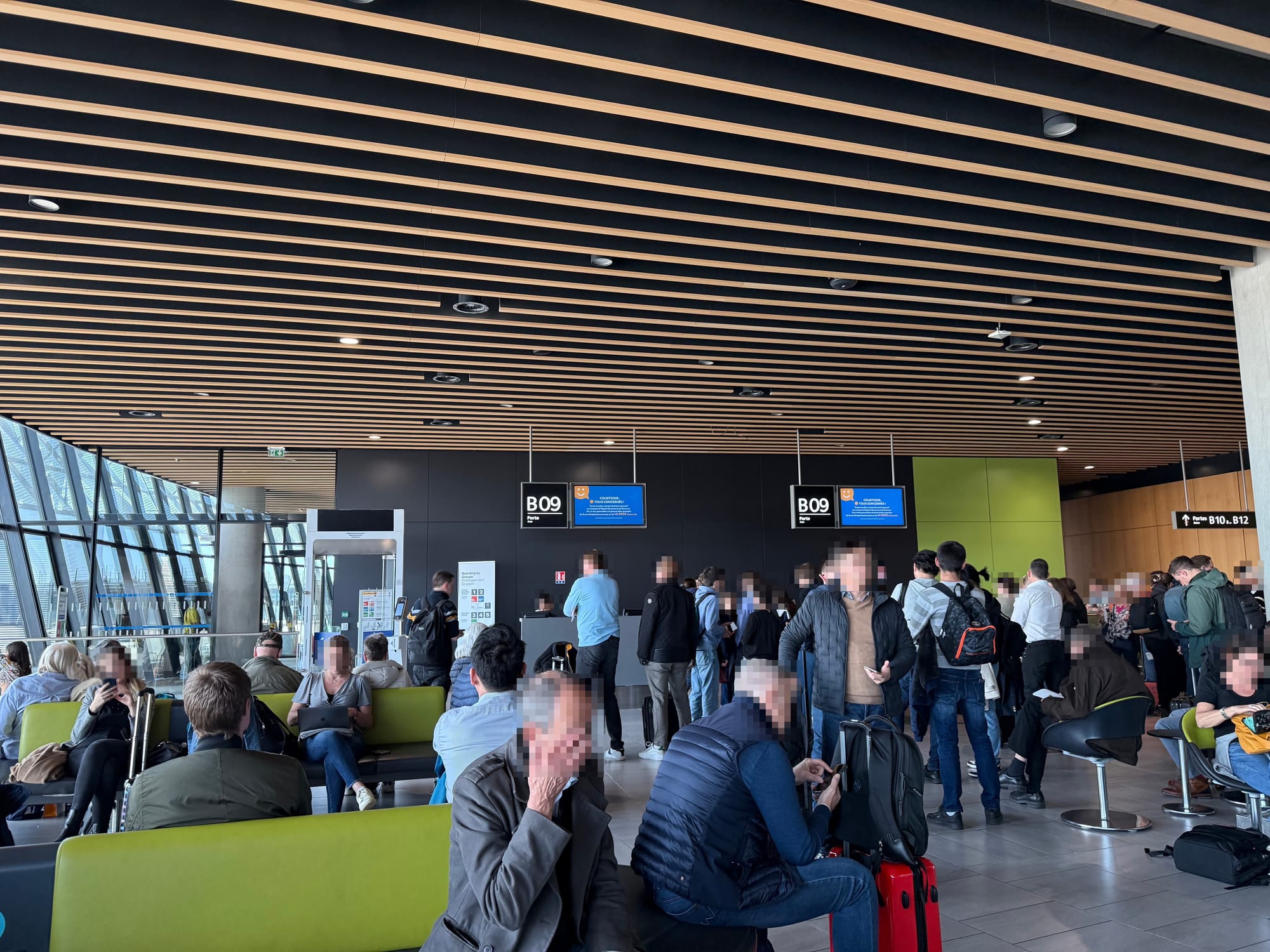 Interior, airport gate area, signed as B09. Wooden slat ceiling with grey stone effect tile floors, and bench seat islands with a lime green covering that's also on one of the walls. Many people mill around the gate area.