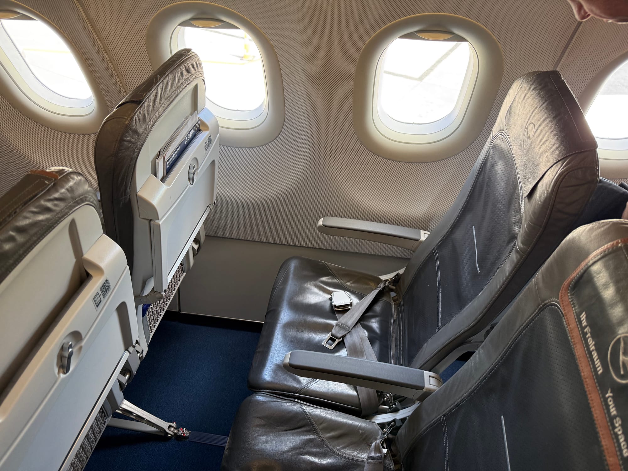 Interior, aircraft cabin, Lufthansa A320 family. A grey leather-covered slimline economy seat (Recaro NEK), with the one in the middle featuring the "Ihr Freiraum/Your Space" leather antimacassar headrest cover with a mid-brown accent.