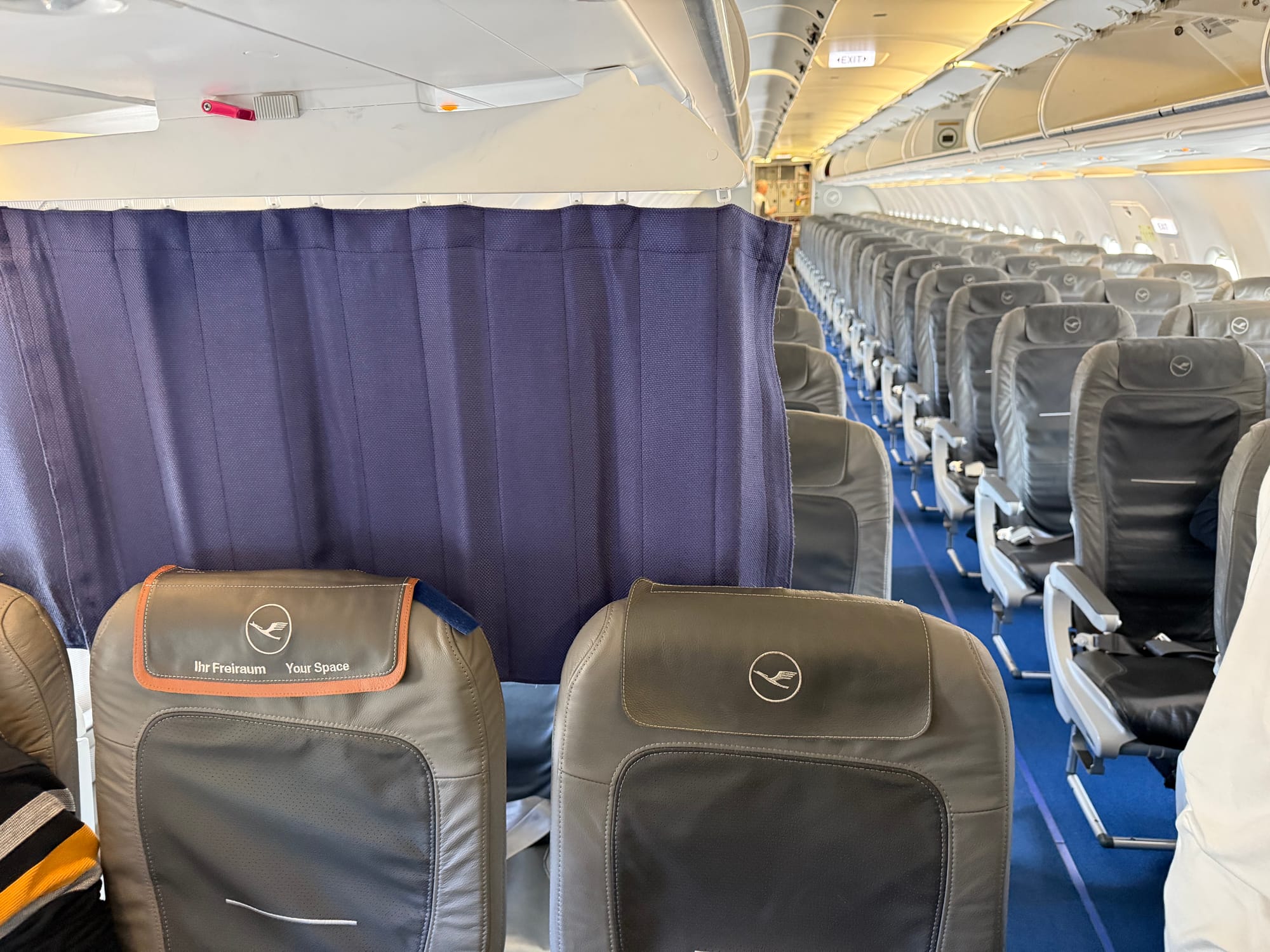 Interior, Lufthansa A320 family aircraft. Looking backwards from the last row of Eurobusiness, with the curtain separating it from the rest of economy, which are the same grey seats but without the central "Ihr Freiraum [Lufthansa logo] Your Space" mid brown-accented headrest cover.