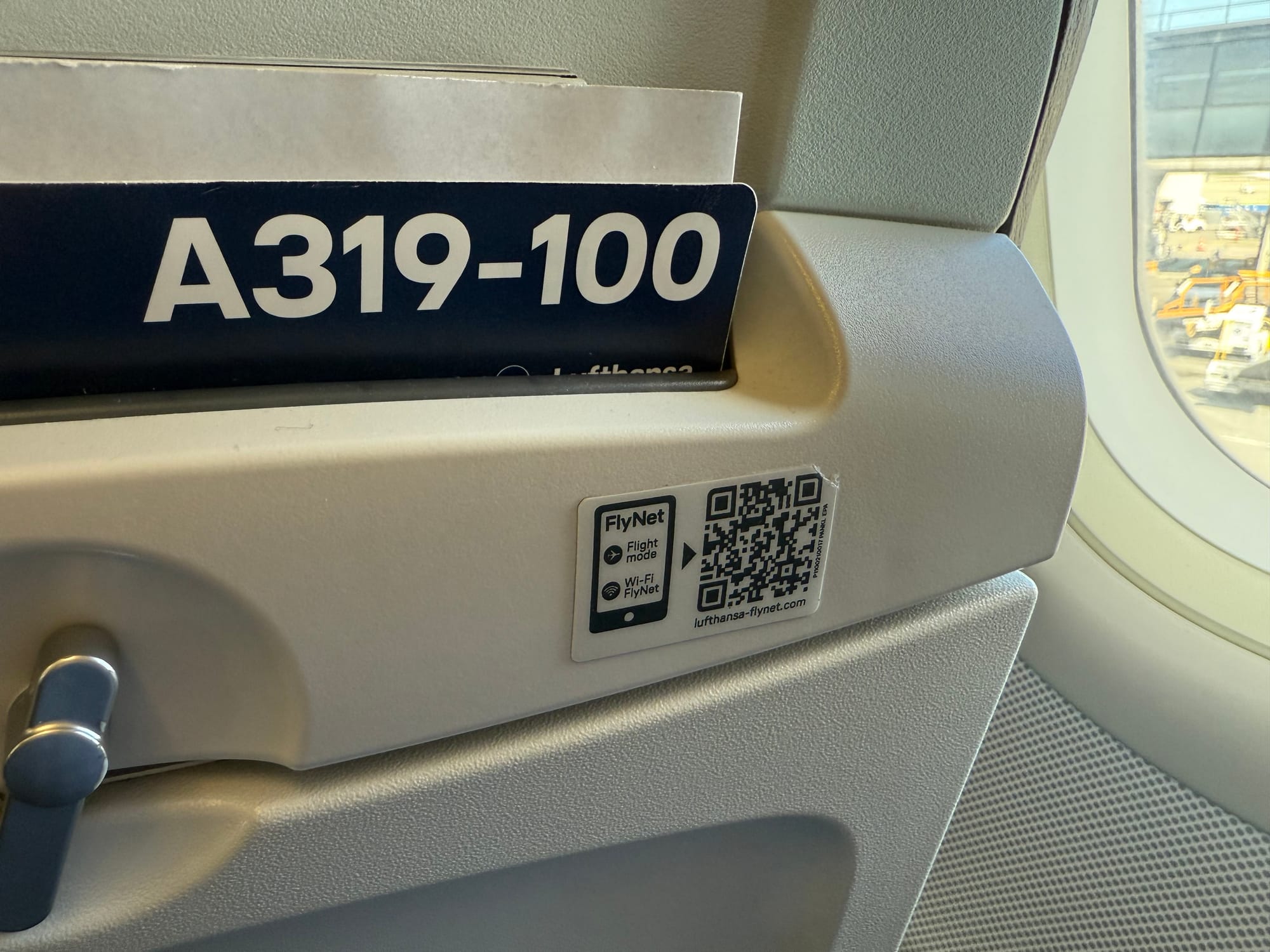 Closeup, airline seatback literature pocket. The A319-100 safety card is visible, as is a QR code for lufthansa-flynet.com with a small tablet logo next to it.