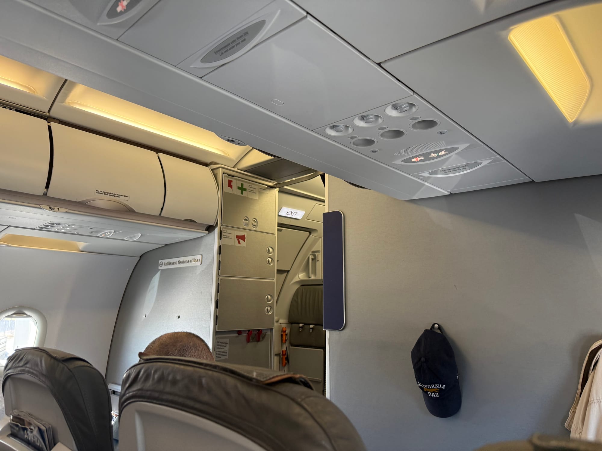 Interior, Lufthansa A320 family aircraft. Looking forward from row 2 to row 1 and the bulkhead, which is an industrial mid grey. A small sign says "Lufthansa Business Class" on the left bulkhead. Also visible are storage cupboards and galley insert slots, in very similar greys. The only colour is a small blue decorative panel on the right hand side. A dark blue hat is hung up on the front bulkhead, with "California Dad" printed on it.