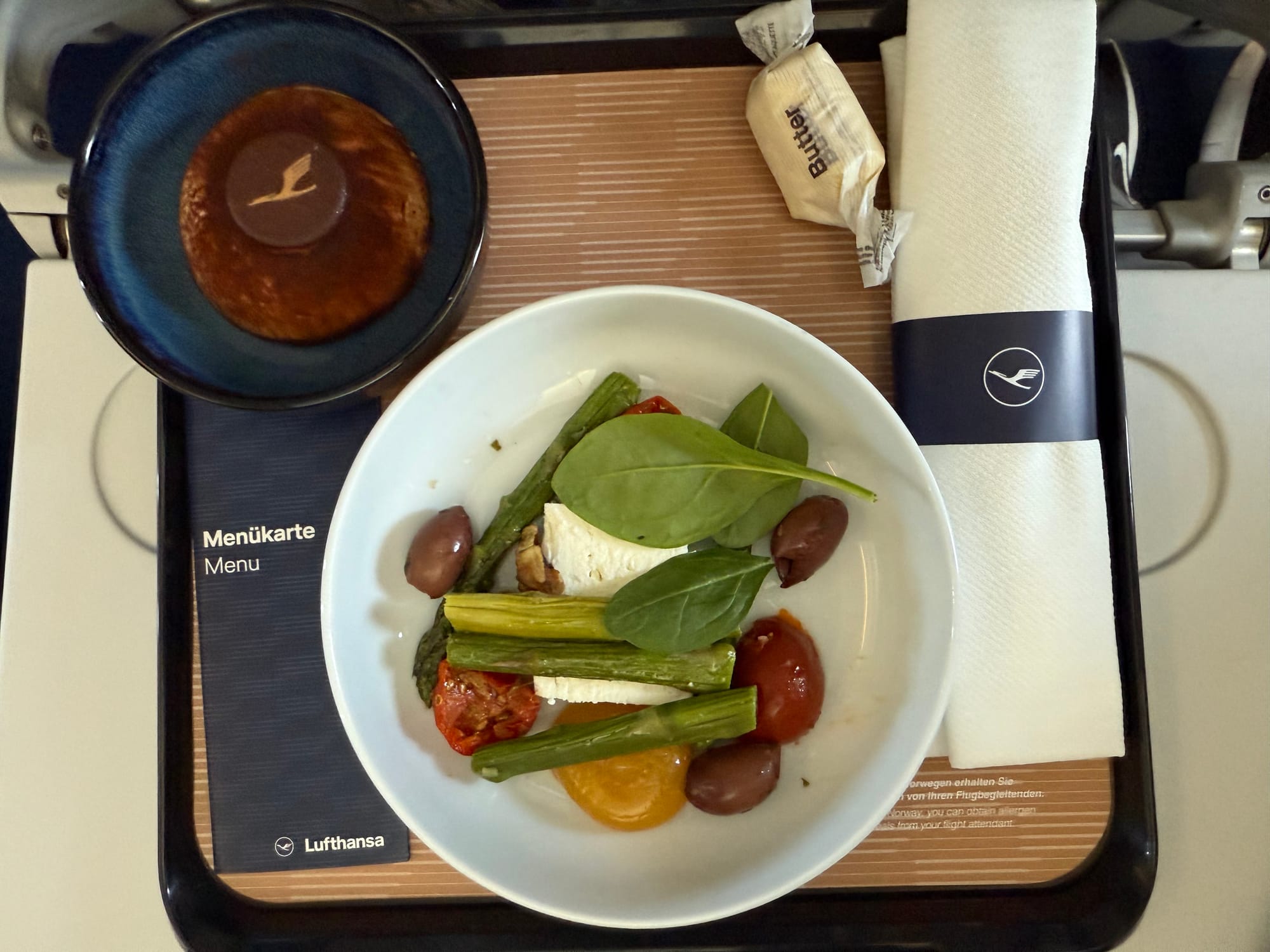 Closeup, airline meal tray on a tray table. A centre white bowl-plate has four pieces of asparagus, two pieces of feta, three pleaves of spinach, three sundried tomatoes and a few olives, plus a piece of walnut. To the top left is a small blue bowl with a chocolate-powder-covered firm dessert dome on it, with a Lufthansa logo chocolate on top. A twist of butter and a napkin that looks like it contains cutlery are on the right, with the menu on the left.