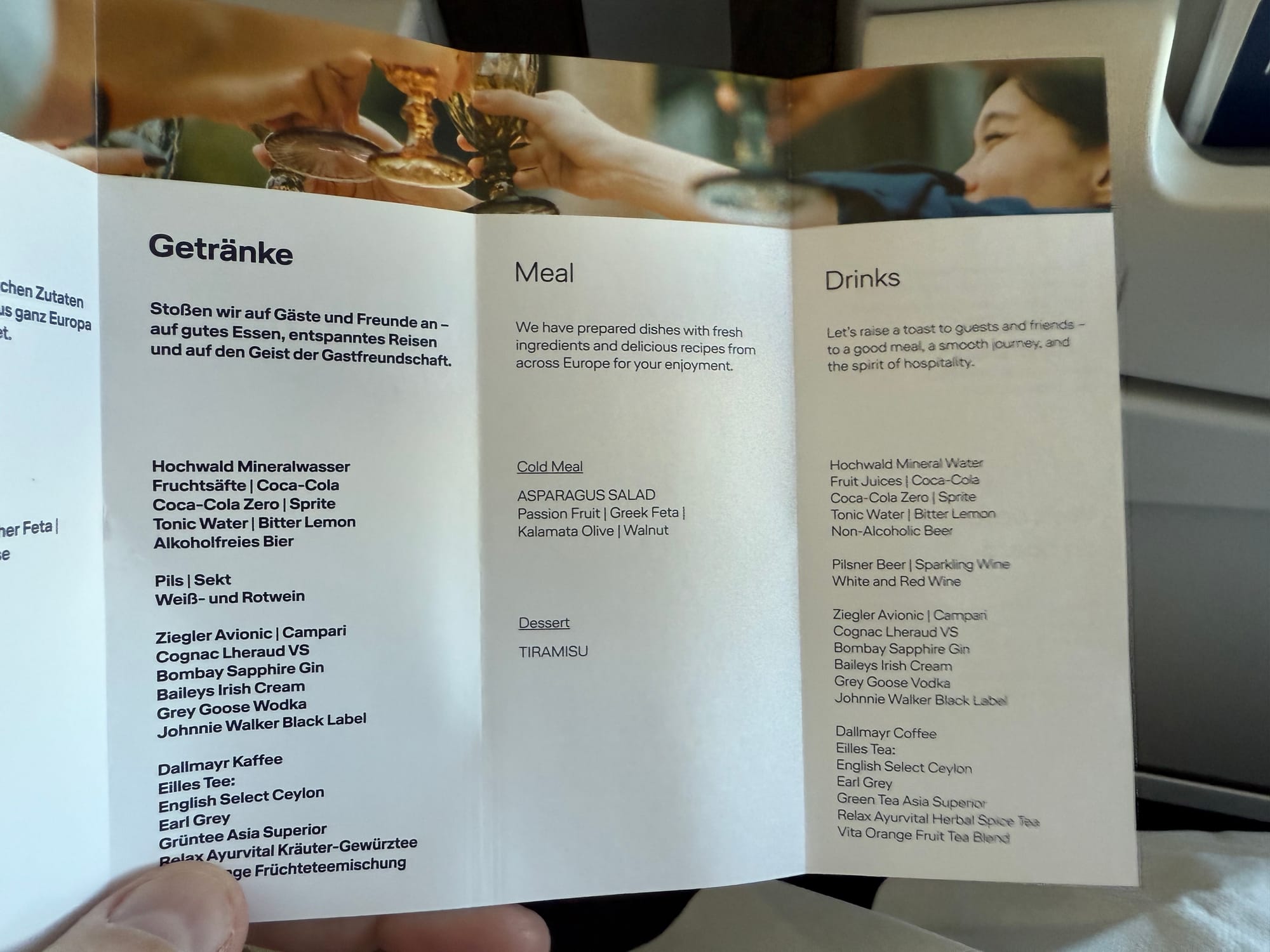 Lufthansa menu in English and German, in total about half the size of a piece of printer paper, then folded into four vertically. The english reads: "We have prepared dishes with fresh ingredients and delicious recipes from across Europe for your enjoyment." The "Cold Meal" is listed as "ASPARAGUS SALAD, Passion Fruit, Greek Feta, Kalamata Olive, Walnut". The "Dessert" is listed as "TIRAMISU". There is a drinks list on the right, with the header "Let's raise a toast to guests and friends — to a good meal, a smooth journey, and the spirit of hospitality". On offer are: Hochwald Mineral Water, Fruit Juices, Coca-Cola, Coca-Cola Zero, Sprite, Tonic Water, Bitter Lemon, Non-Alcoholic Beer, then Pilsner Beer, sparkling Wine, White and Red Wine, then Ziegler Avionic, Campari, Cognac Lheraud VS, Bombay Sapphire Gin, Baileys Irish Cream, Grey Goose Vodka, Johnnie Walker Black Label, then Dallmayr Coffee, Eilles Tea: English Select Ceylon, Earl Grey, Green Tea Asia Superior, Relax Ayurvital Herbal Spice Tea, Vita Orange Fruit Tea blend.