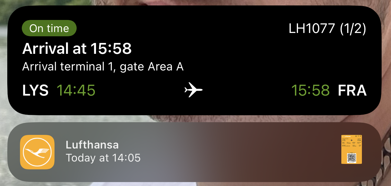 Screenshot of an iPhone lock screen with two notifications. The first is a Live Activities notification showing: On Time flight, LH1077 1/2, Arrival at 15:58, Arrival terminal 1, gate area A, LYS 14:45 [plane icon] 15:58 FRA. The second is a boarding pass notification with the Lufthansa logo in white on a yellow background, the word "Lufthansa", "Today at 14:05", and a thumbnail of a boarding pass.
