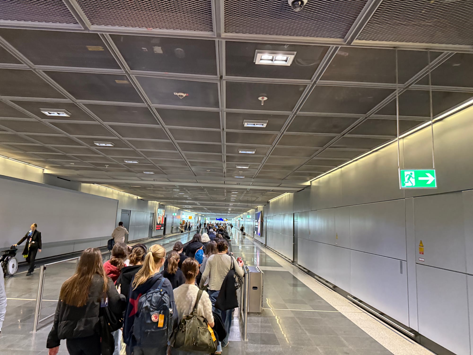 Interior, Frankfurt Airport corridor. A crowd of people are approaching and walking on a moving walkway. Everything else is an ugly industrial grey: the grates on the ceiling, the cladded walls, the worn tile floors. The lighting is industrial grim.