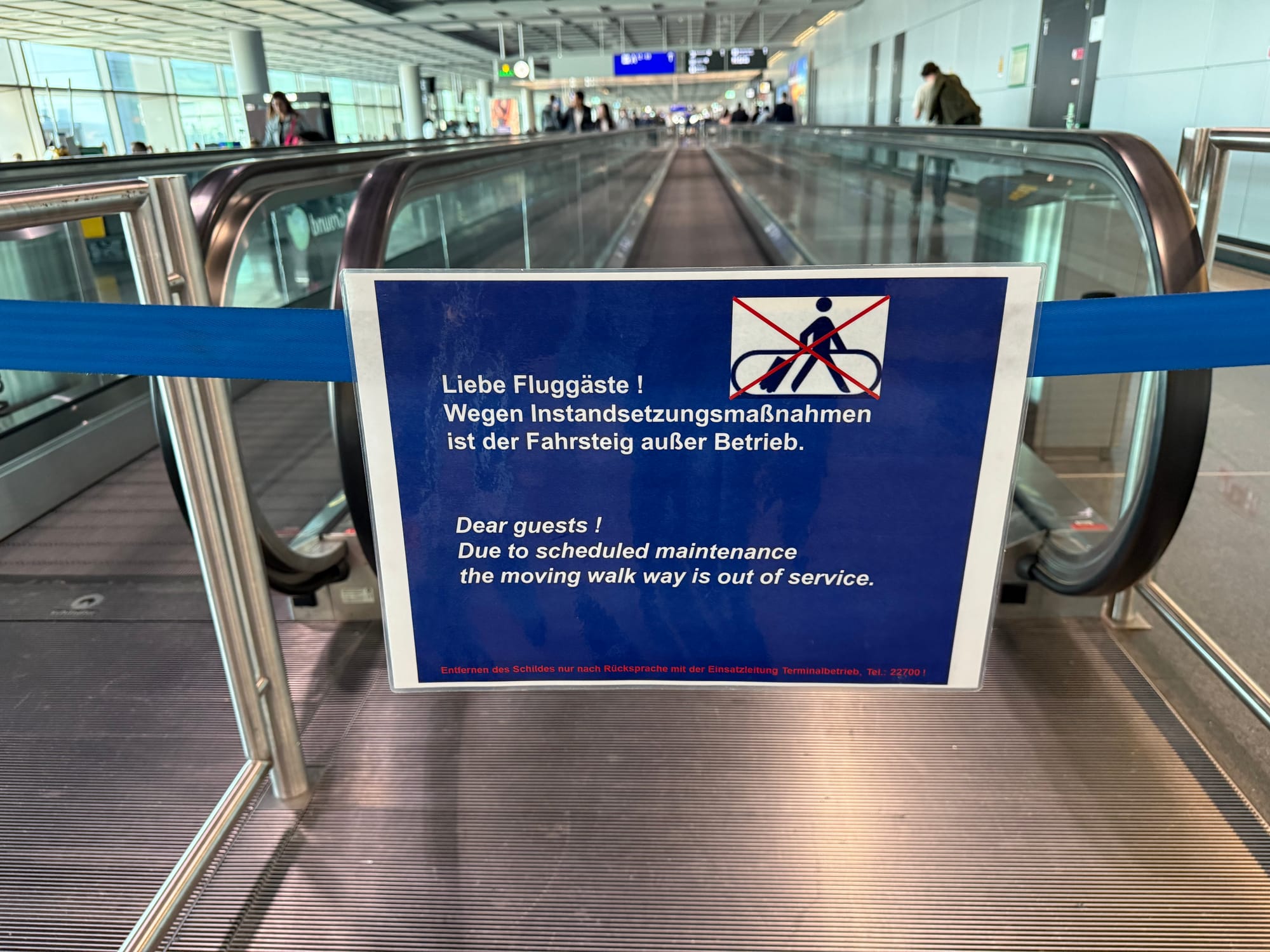 Interior, very long airport corridor (it is Frankfurt). A sign in English and German blocks off the entry to a moving walkway "Dear guests ! Due to scheduled maintenance the moving walk way is out of service". There is no visible maintenance taking place.