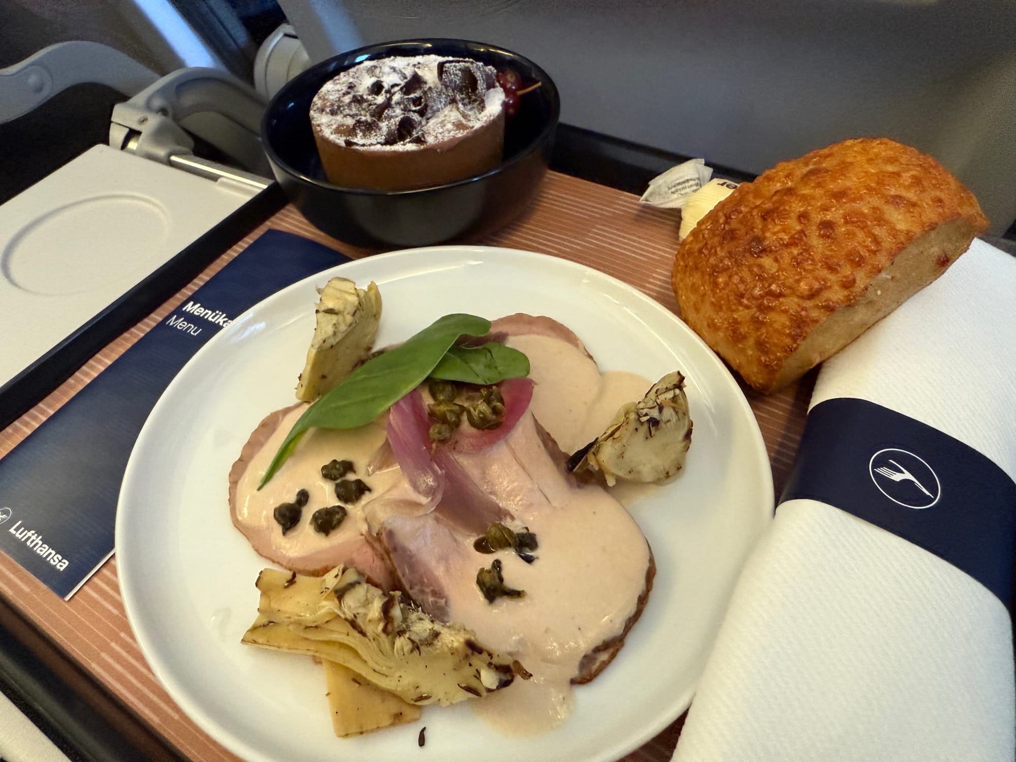 The same meal tray as before, but now there are three slieces of veal with a pink-beige sauce, three pieces of roasted asparagus, a few capers on each and some spinach leaves on top. A bread roll is on the right, and the dessert looks like a firm chocolate mousse timbale.