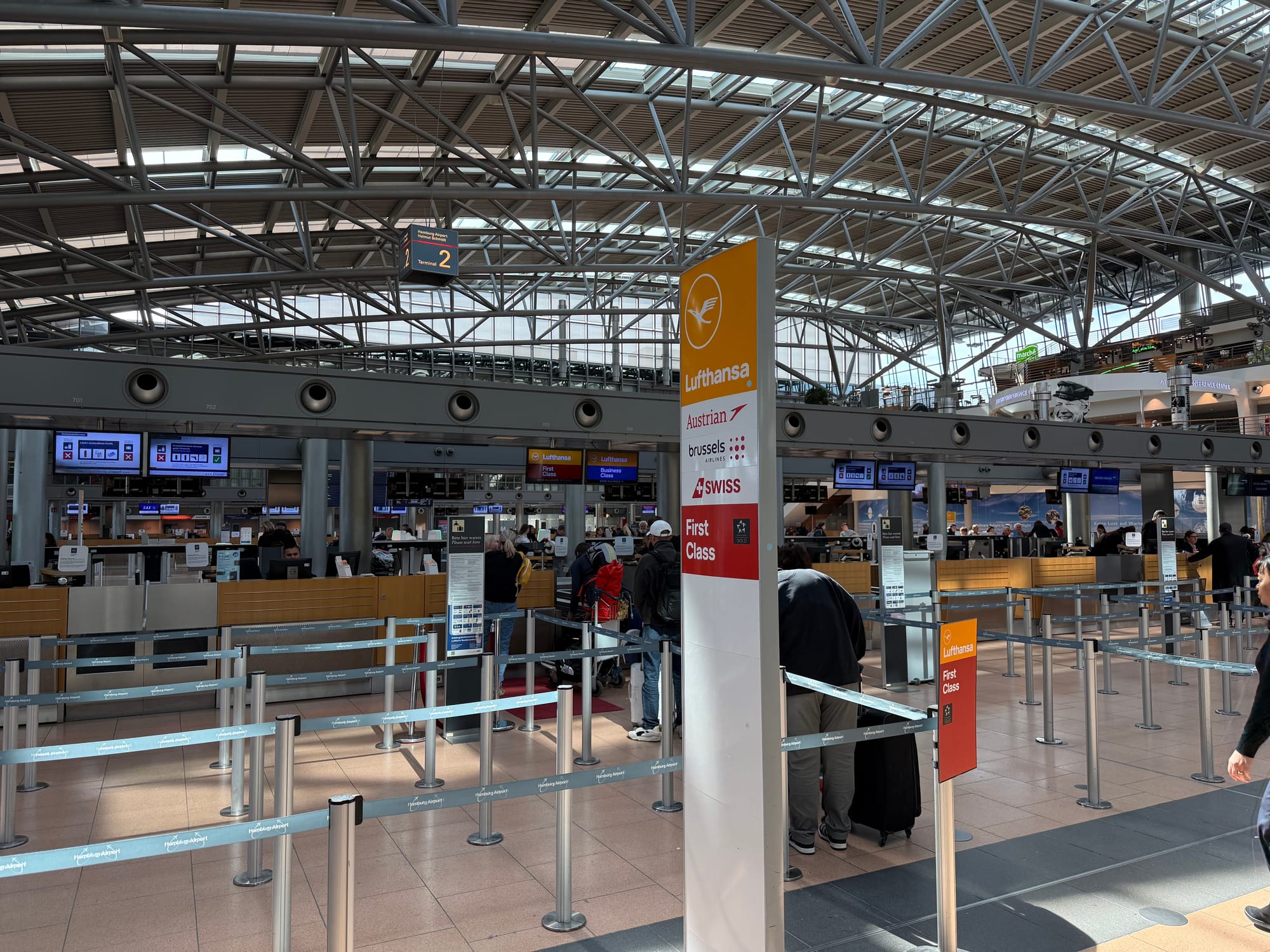 Interior, airport checkin hall, Lufthansa (and Austrian, Brussels and Swiss) desks. One first class and one business class desk are operational, with several desks empty. A queue of about five people has formed..