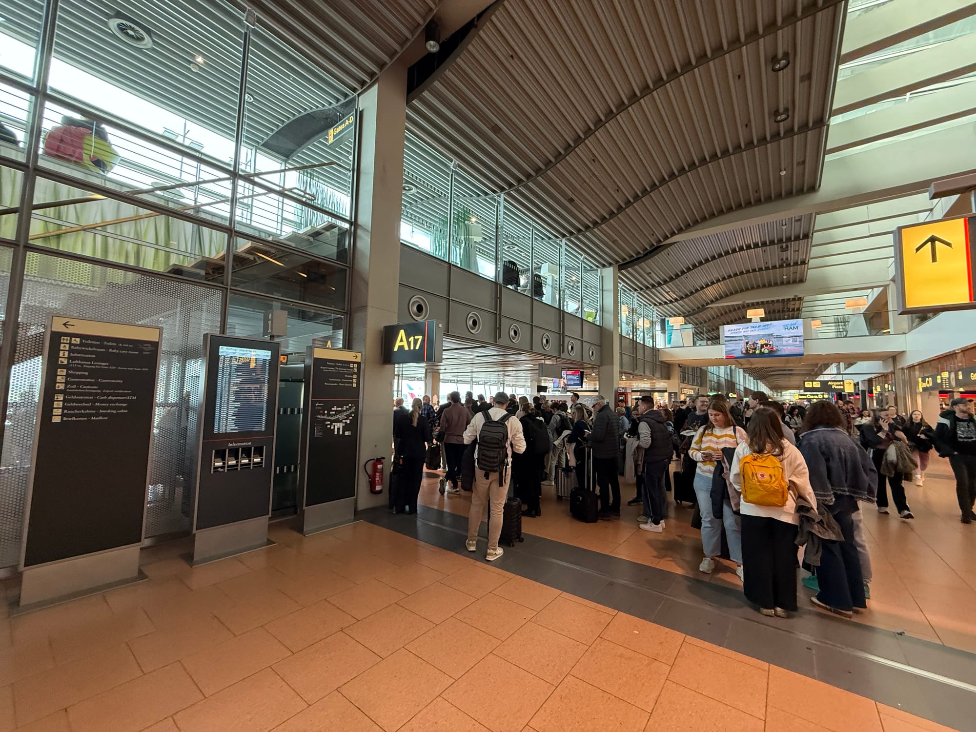 A very crowded boarding gate area (it is Hamburg Airport), with passengers crowding across about 3/4 of the corridor walkway space and blocking others from getting past.