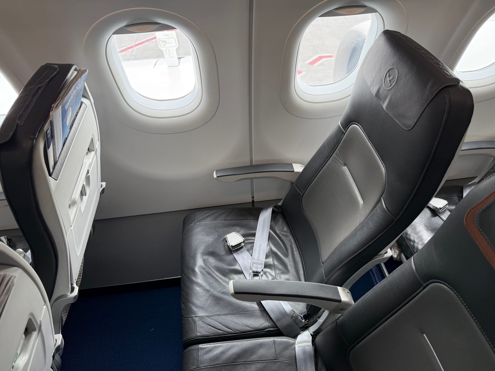 Interior, aircraft cabin, Lufthansa A320 family. A grey leather-covered slimline economy seat (Geven Essenza), looking a little more padded and comfortable than the previous one, with the seat in the middle featuring the same "Ihr Freiraum/Your Space" leather antimacassar headrest cover with a mid-brown accent.
