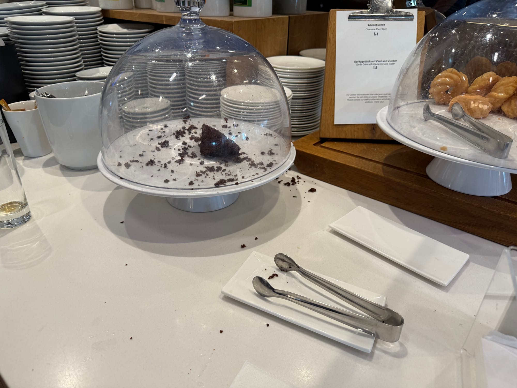 Interior, airline lounge. A sad single piece of chocolate cake sits under a glass fly dome, with crumbs all over both the cake stand and the sufrace beneath it. There is also a stand of what appear to be glazed cruller-style donus.