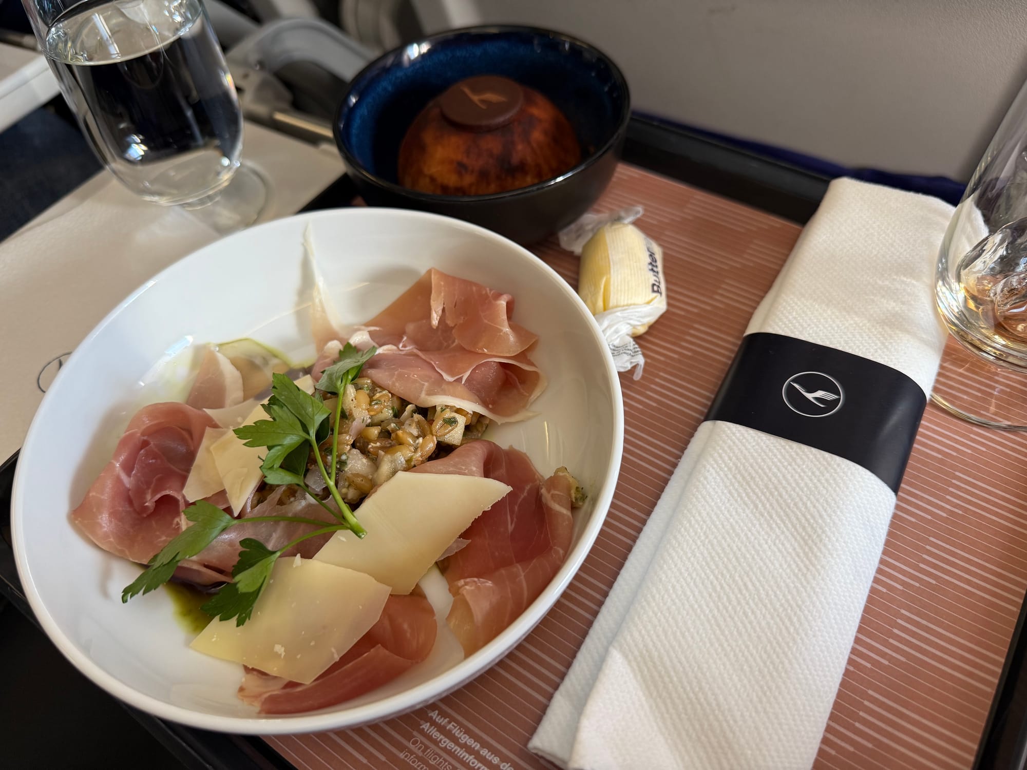 The same meal tray layout as before, only with a main bowl-plate of four pieces of prosciutto-style ham, a few shavings of parmesan-style cheese, and a small prepared salad that looks like grains and perhaps small cubes of potato.