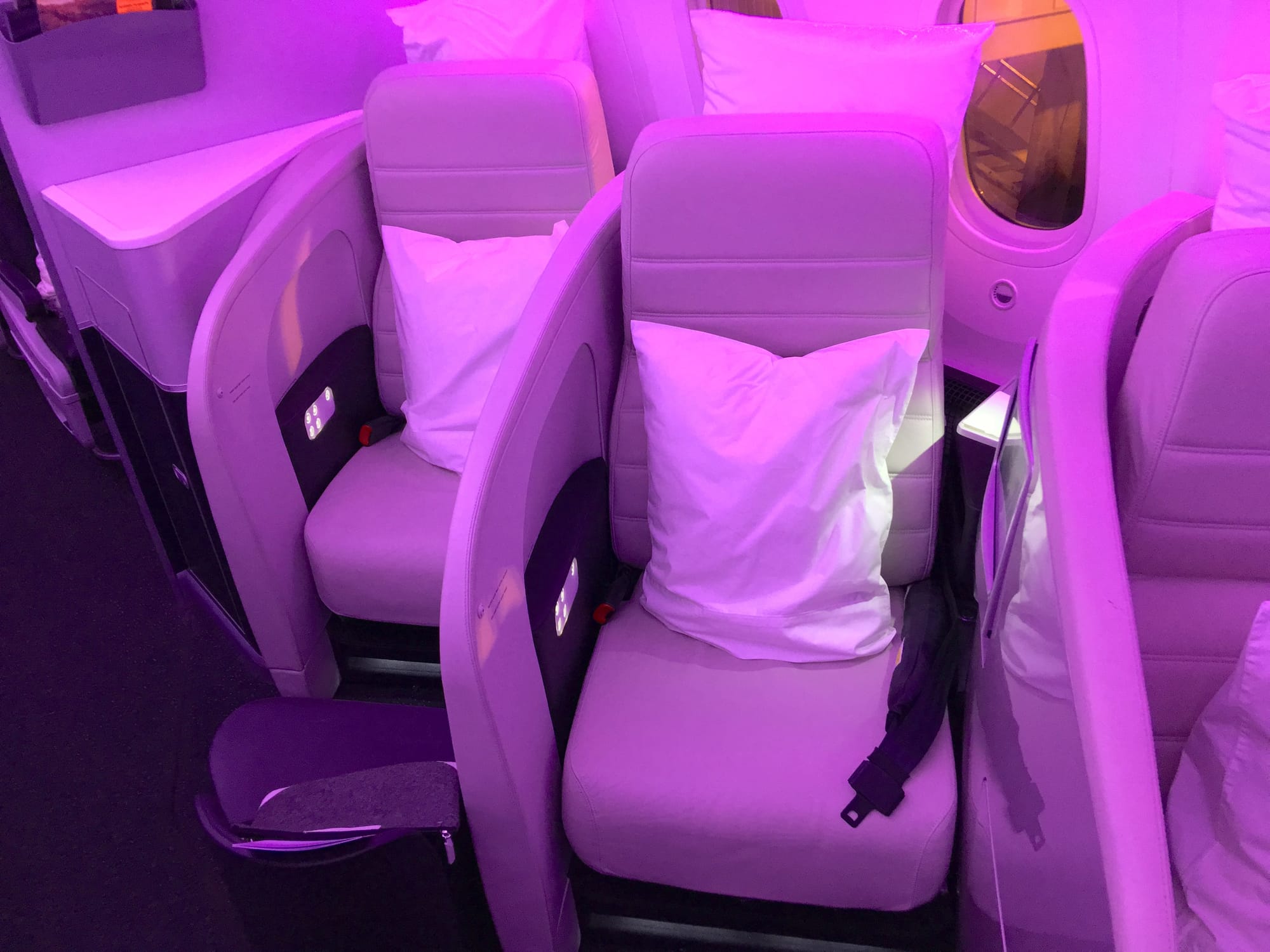 Interior, airline cabin, lots of pink LED light wash, large 787 signature windows. An off-white seat is facing the camera.