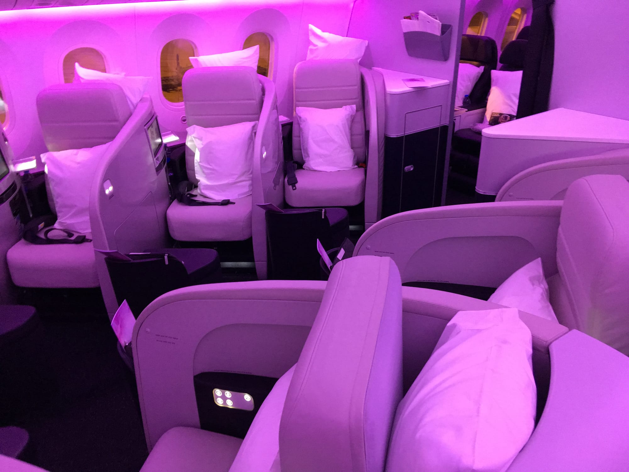 Interior, airline cabin, night, with magenta LED light wash. Inward-facing herringbone seats in an off-white colour catch the light.