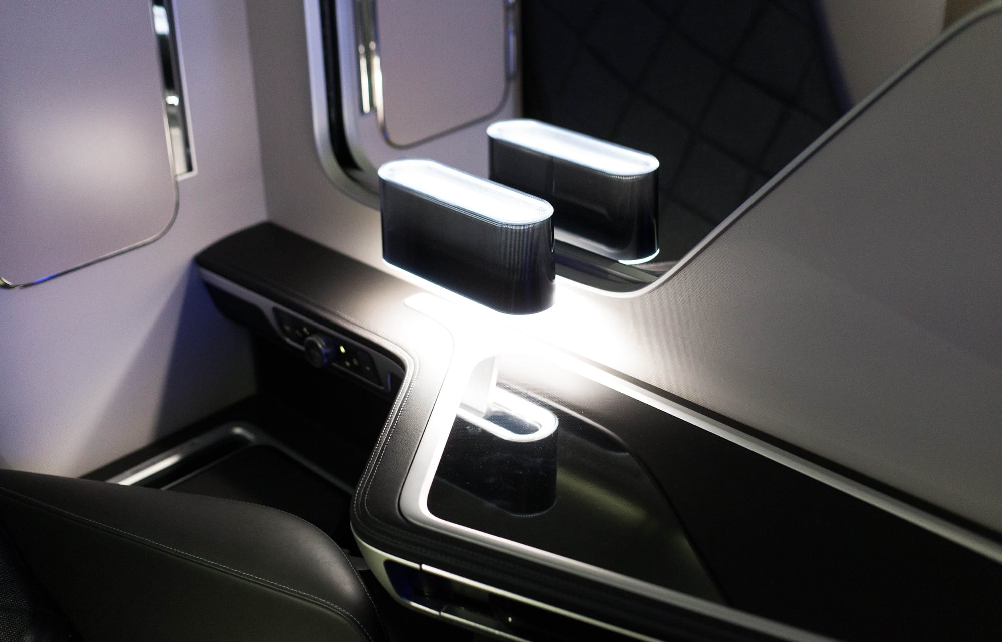 Closeup, airline seat elements, centre pair of two first class seats. A blue-white oval lozenge-shaped lamp shines brightly onto a reflective black side table surface, so brightly that its support arm is only visible in the reflection. Behind and below it is a control panel with a twisting knob. The walls are white thermoplastics with aluminium-effect metallic edging, also seen elsewhere on the closeup. Black leather is on the armrest and the table surrounds. The overall aesthetic is very automotive.