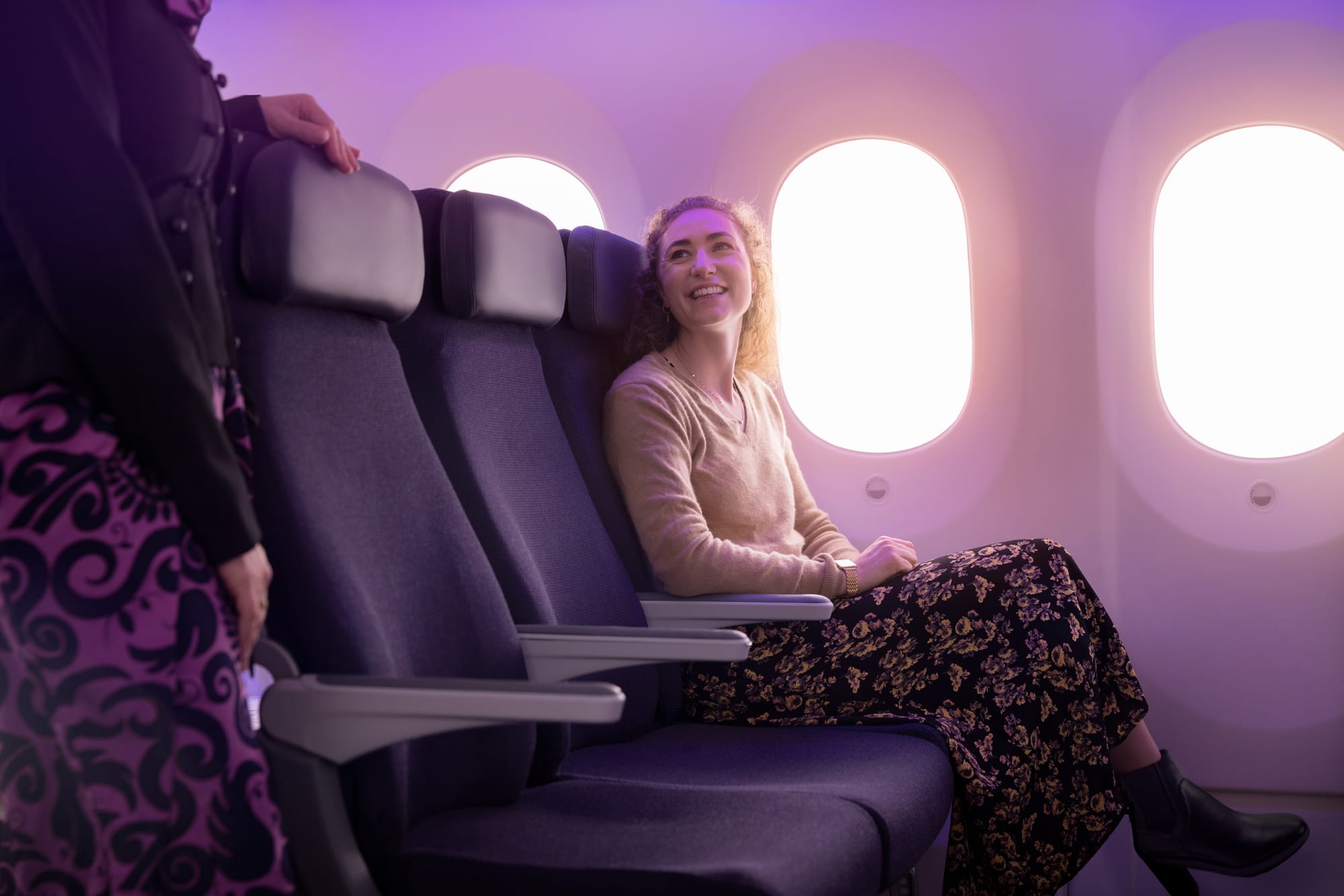 Interior, airline cabin, economy. A woman smiles up at a flight attendant half out of shot. The seat is sculptured and contoured, with a headrest, and looks well-padded. The seat fabric is in black or near-black.