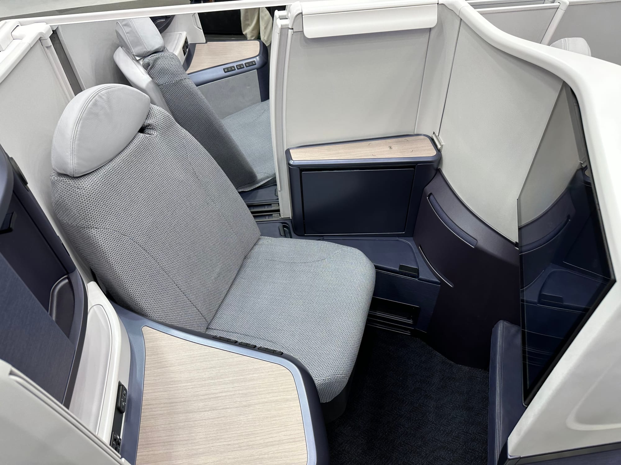 A seat mockup at a trade show. It is a business class herringbone angled towards the camera. There's a lot of grey thermoplastic above the seat pan level and a lot of dark blue thermoplastic below it. The seat itself is very grey. The overall aesthetic feels quite dated and not especially luxurious.
