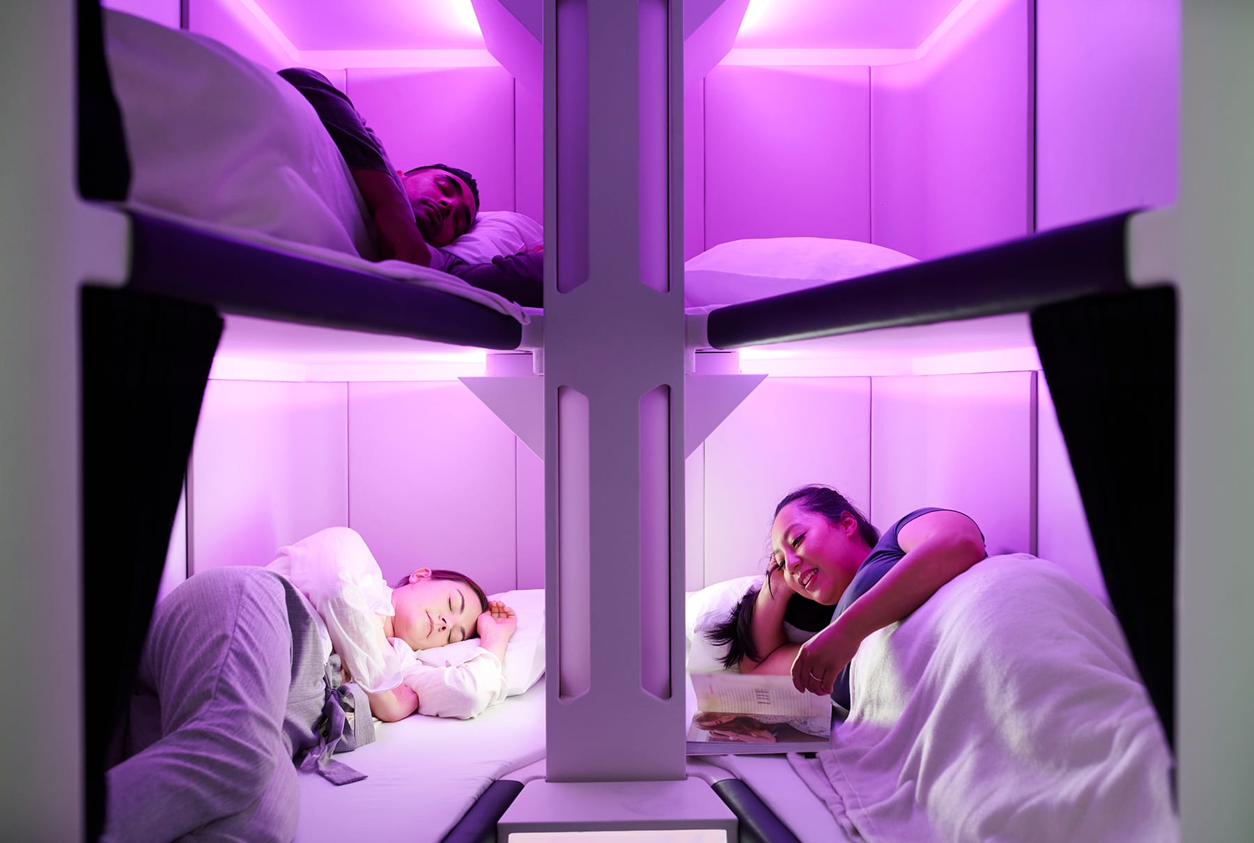 Interior airline cabin, lit with pink LEDs. We see four train couchette-style bunks with people sleeping on them.