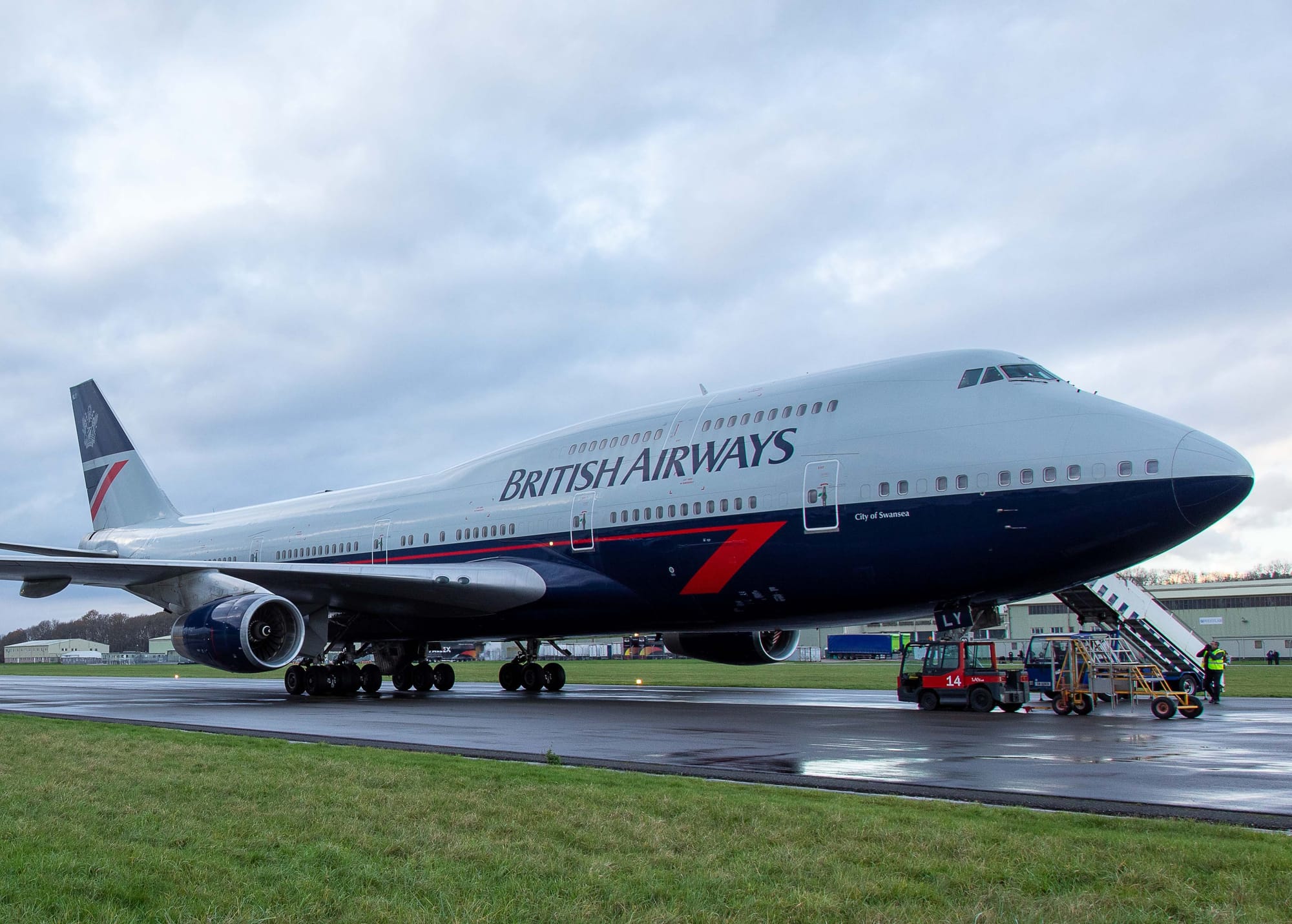 A BA 747-400 sits on the runway with ground vehicles nearby. Its livery is white on the top, dark blue from just underneath the windows, with a thin red cheatline extending from the rear of the aircraft all the way to just before the front doors, at which point the cheatline gets thicker (like a calligraphy nib) and descends at about a 45° angle.