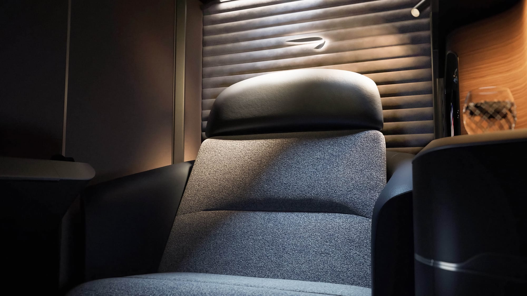 Seat close up, interior suite, looking up at the seat from about knee height. The light is playing on the padded, quilted velvet-effect rear wall with an internally lit BA ribbon speedmarque brand element. There is a very tactile look to the medium grey seat fabric, the warp and weft of which give a 3D effect.