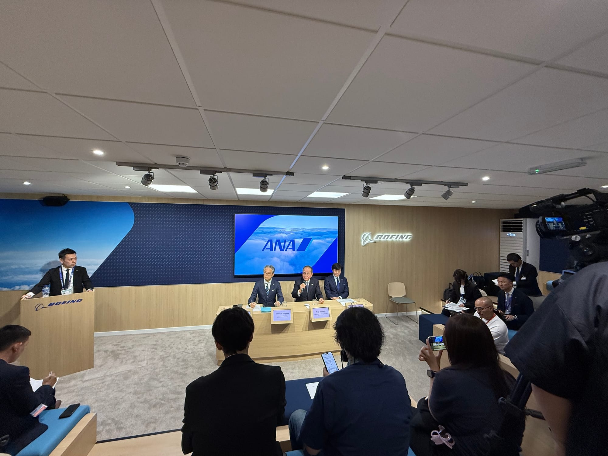 A small press room with risers and the Boeing logo, a trio of execs at a table (with ANA branding on a screen behind), and a further exec at a lectern.