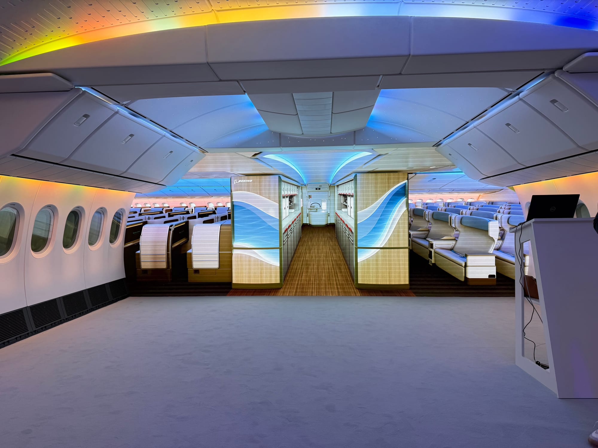 Interior, Boeing 777X cabin mockup. On the right is a lectern with laptop, ahead is a big screen showing a rendering of the view entering from doors 2. A wood and waves effect pair of walls bracket the galley.