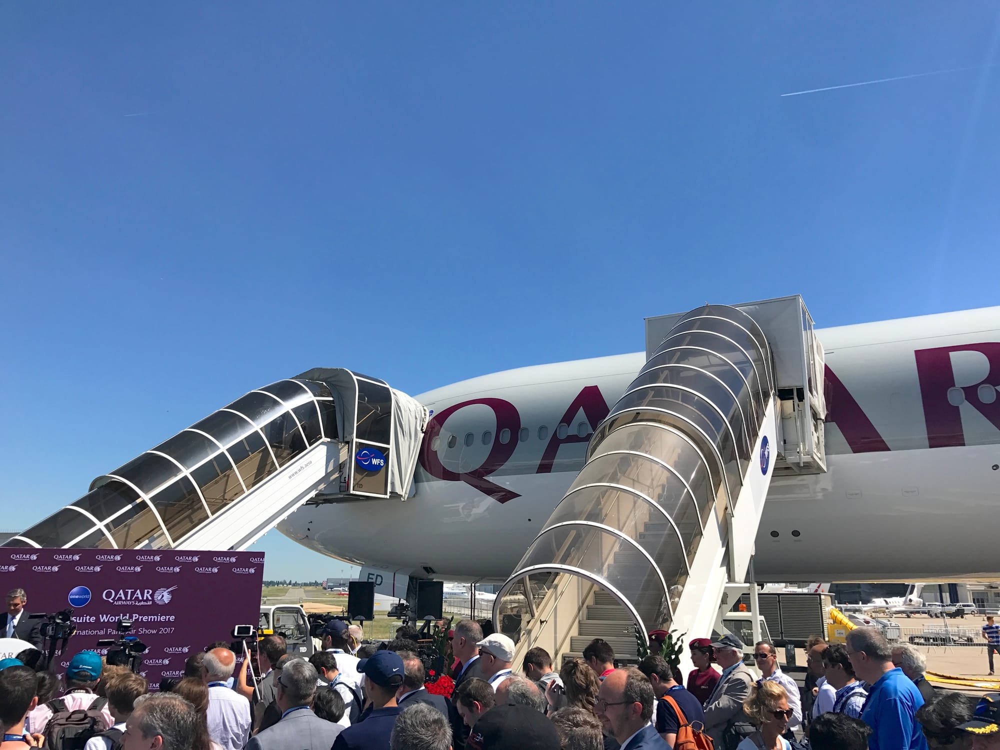 A crowd standing under two airstairs up to a Qatar 777-300ER with the nose registration plates "ED". A Qatar Airways podium is in front of the front airstair.