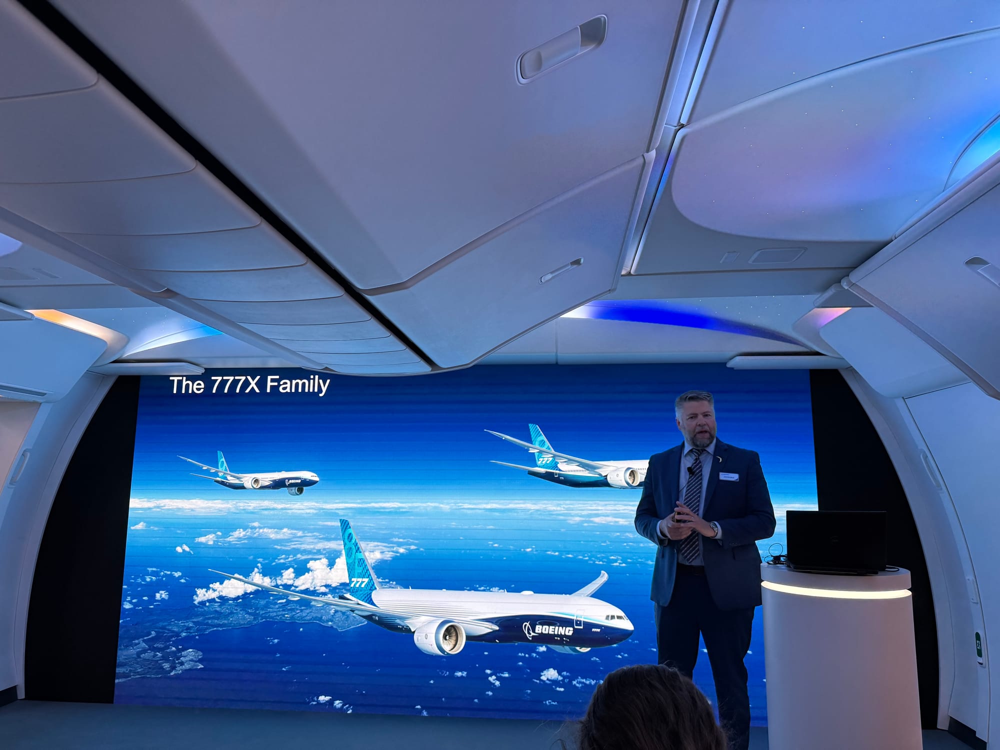 A man stands inside a cabin mockup in front of a screen with a slide: "the 777X family" and three 777X airplanes.