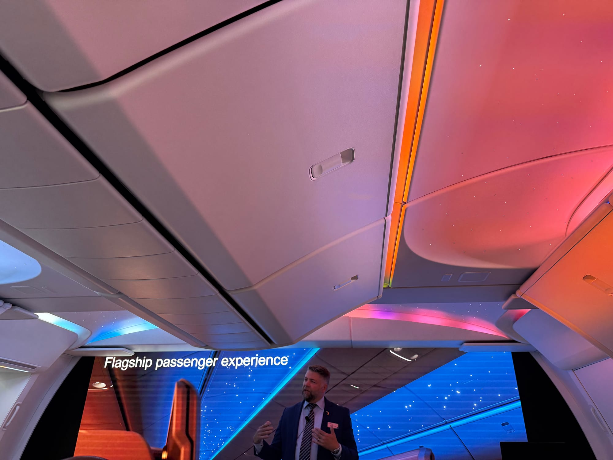 A man standing inside the 777X cabin mockup with multicolour LEDs, in front of a "flagship passenger experience" sign.