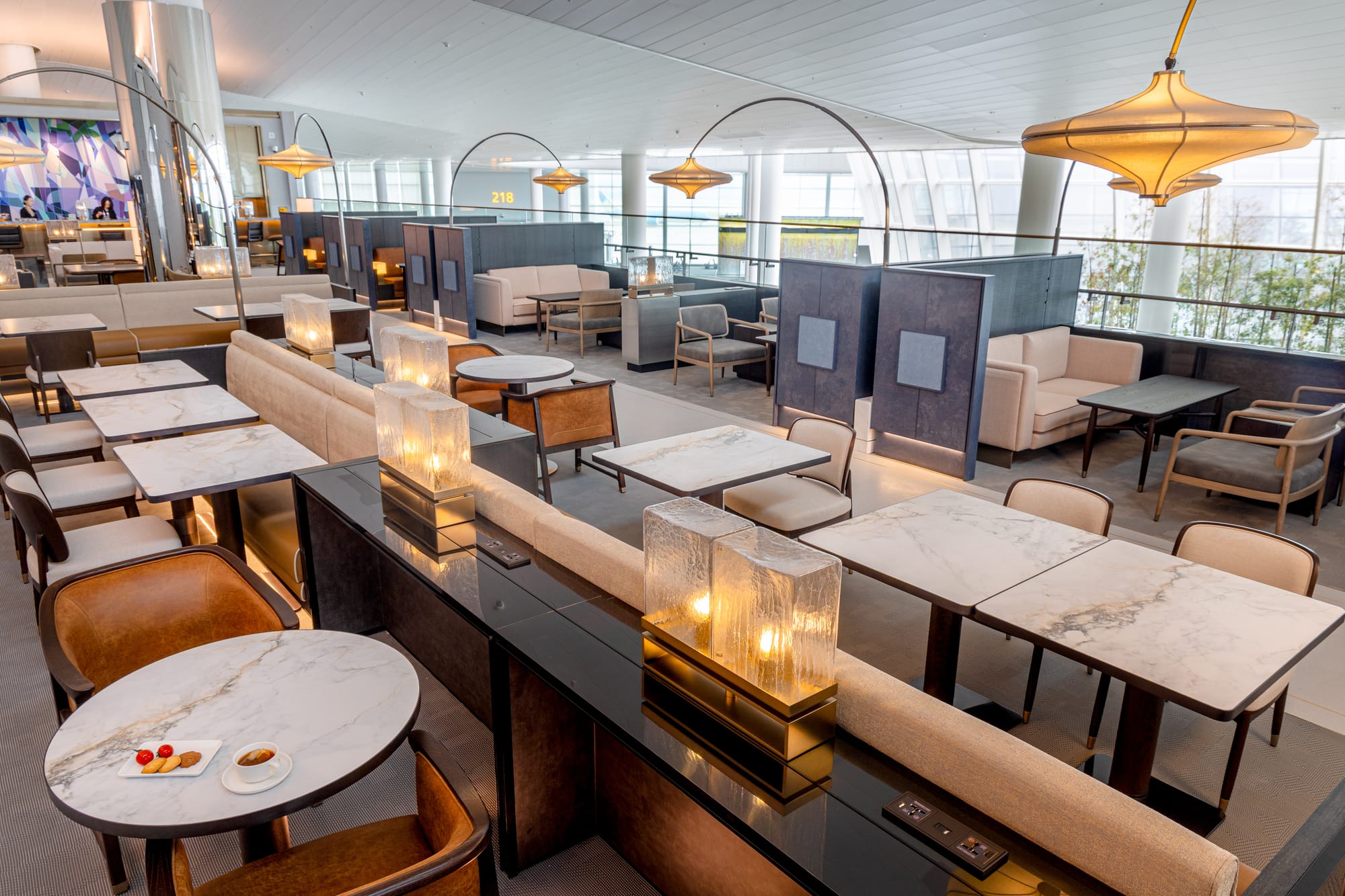 Interior, airline lounge, upper level of a terminal, open to the rest of the terminal. A variety of seats, sofas and tables, some divided by blue or beige dividers of various heights and materials.