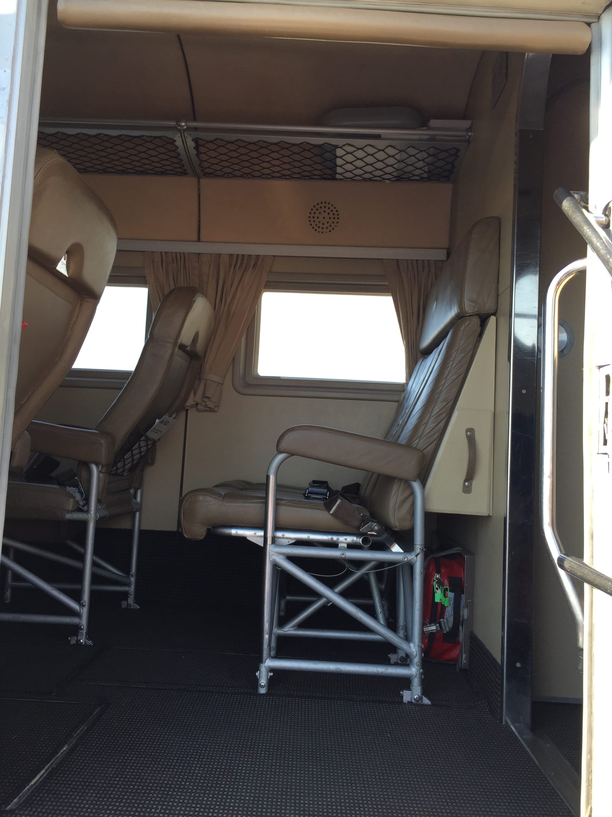 Interior of Tante Ju, with the rear twin pair of seats. Everything is beige apart from a grey carpet, silver metal structures under the seats, and the black overhead netting of the metallic hatracks. The large rectangular windows are framed by curtains and there is a speaker grille in the panel above them.