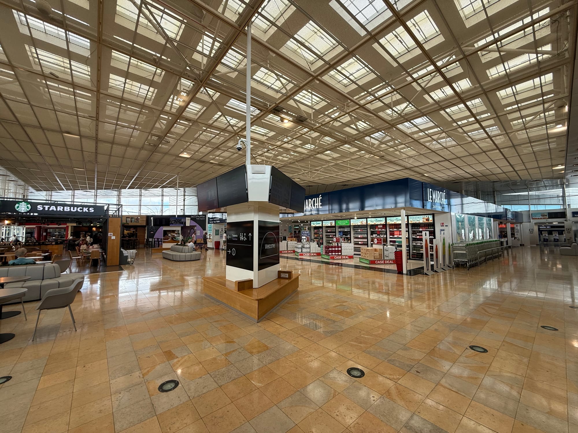 Interior, terminal. Industrial roof, beige and grey tiled floor, and a large duty free in the middle. A Starbucks is on the left.