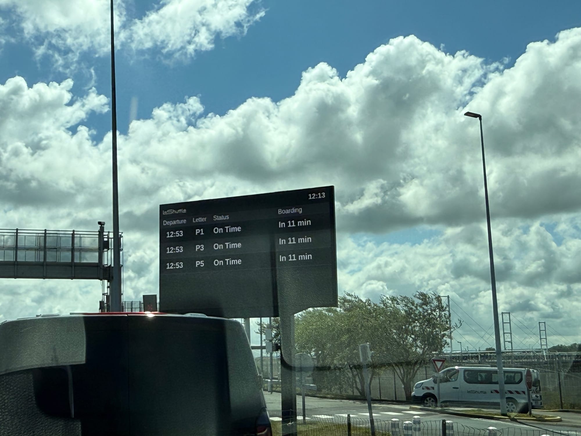 A departures display board in a car park: 12:53 departure, Letters P1, P3, P5. On Time, Boarding in 11 min. Current time: 12:13.
