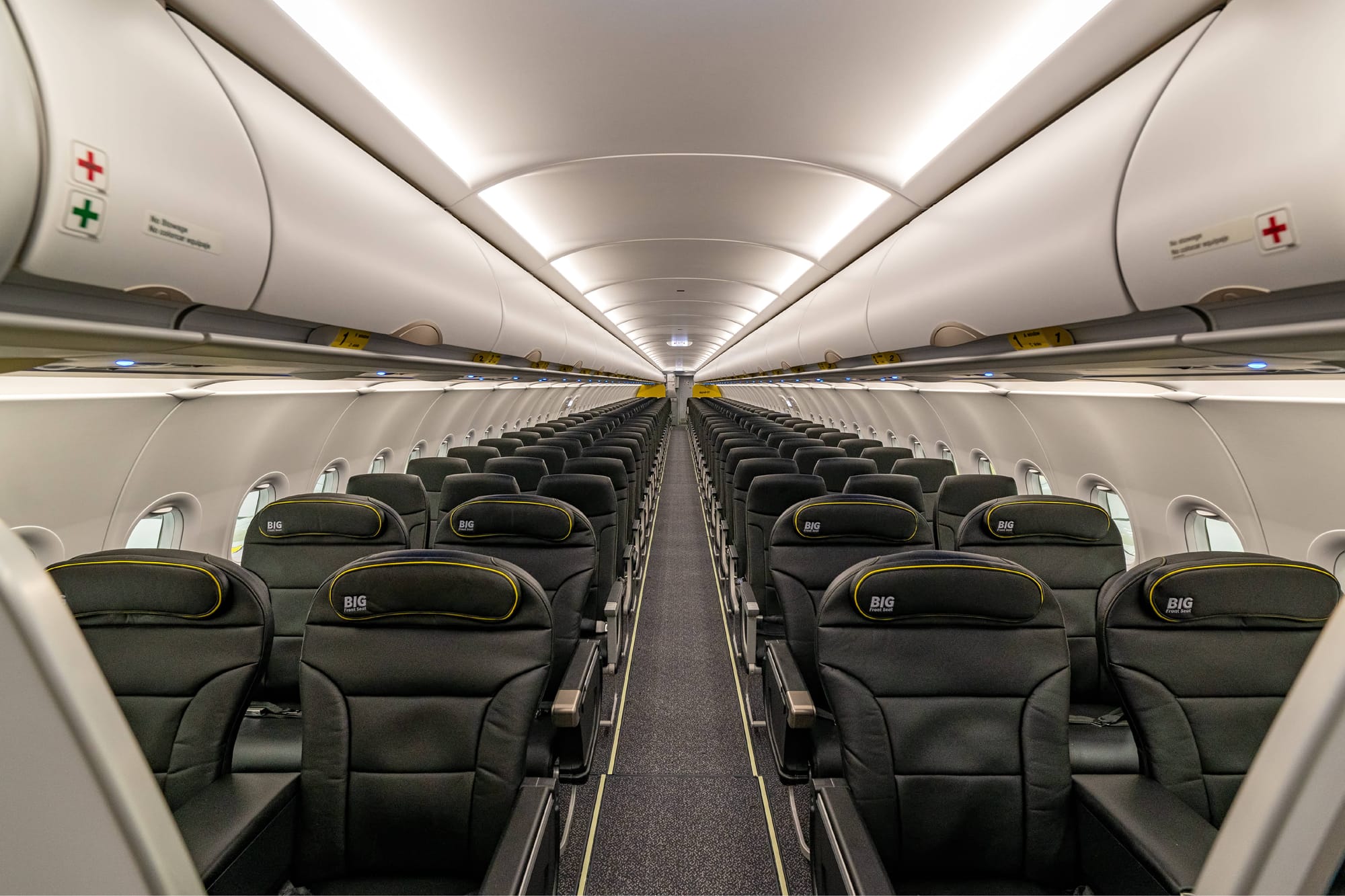 Interior, A320 family aircraft, looking down the aisle from the front of the plane. 2 rows of 2-2 recliners then a lot of rows of regular seats, all in black leather/leather effect.