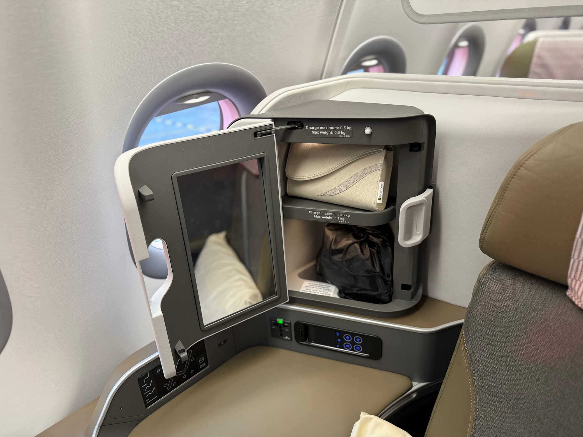 Interior, business class seat, focus on the side console by the shoulder with the cabinet. Inside is a beige amenity kit and a small squashy black sack that looks like it might contain headphones.