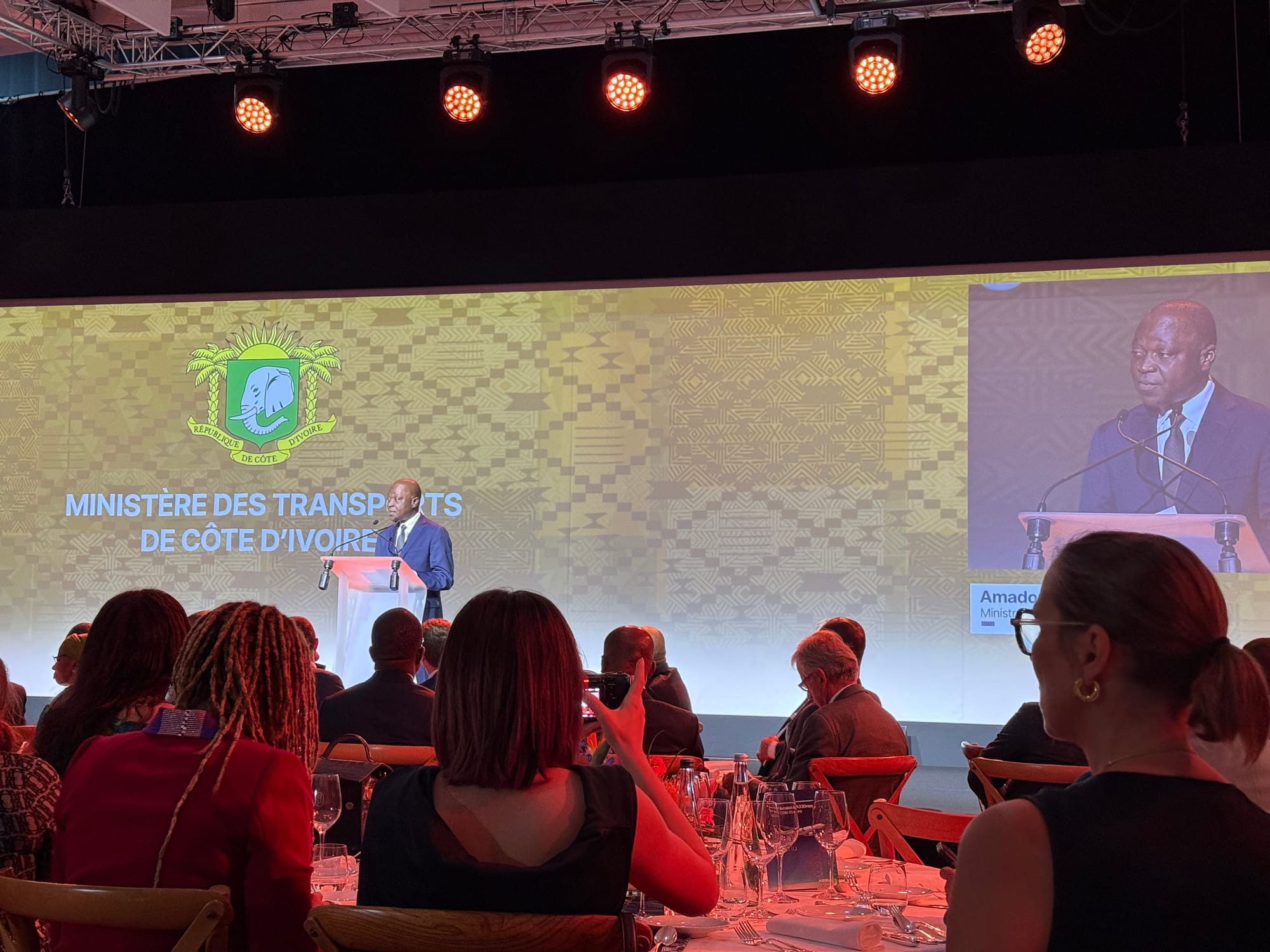 Gala dinner, with a man in a suit speaking in front of a projected sign with the Côte d’Ivoire crest and "MINISTERE DES TRANSPORTS DE COTE D'IVOIRE". His name is on an inset, right, but it is cut off by an attendee's head.