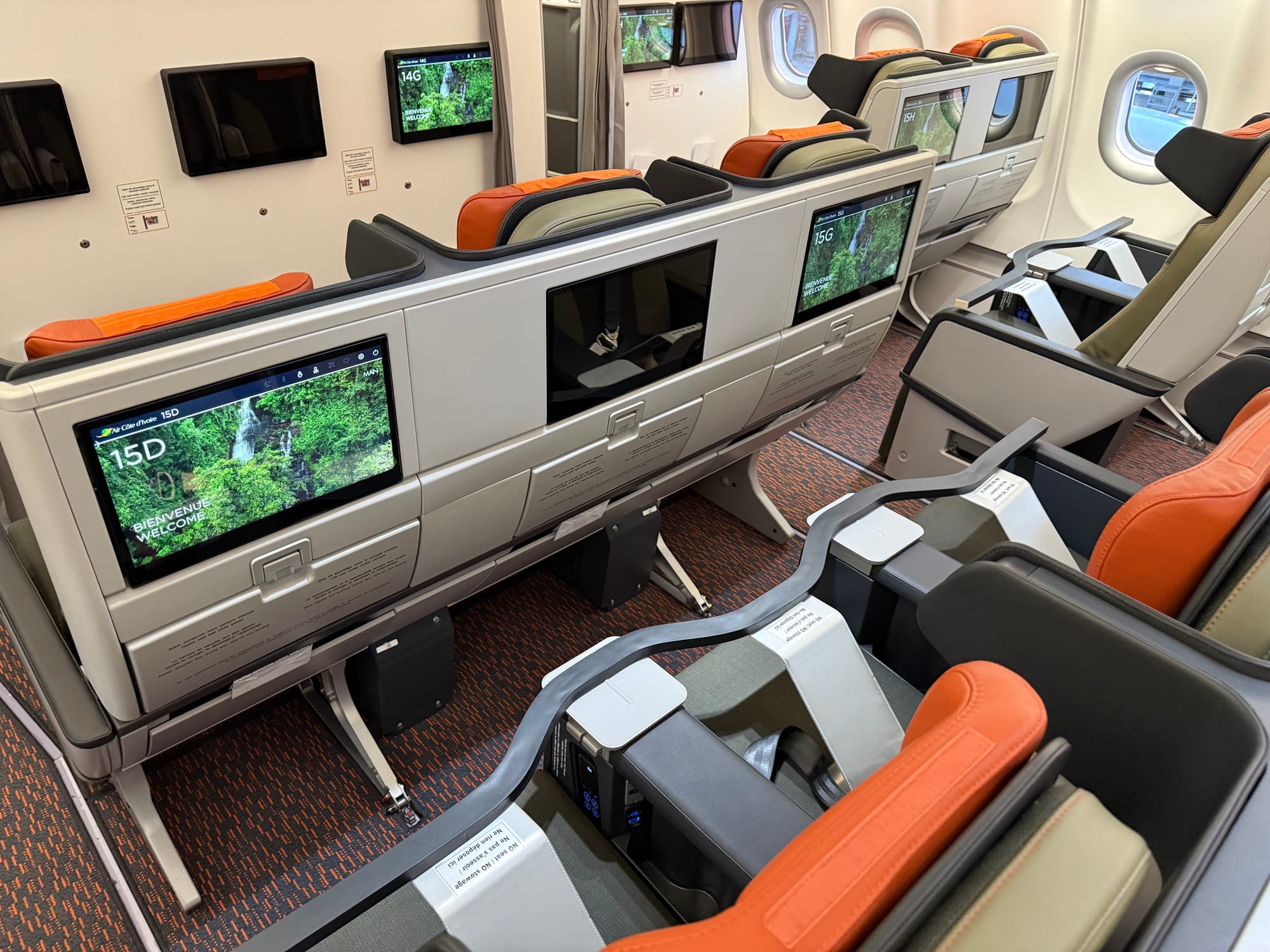 Interior, premium economy class cabin, 2-3-2 layout. Fixed seatbacks in grey with large screens, green seat covers, orange headrests. Row 2 of the 2 visible is blocked off with a bar that says "no seat no stowage" in English and French.