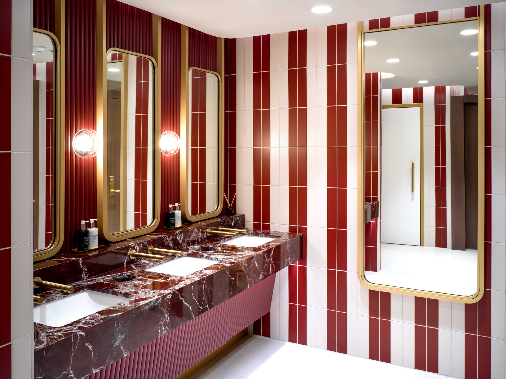 Interior, lounge bathroom, art deco/roaring twenties aesthetic. Red and white striped tiled walls, red-striped walls behind golden mirrors, red marble-effect sinks, red under-vanity space.