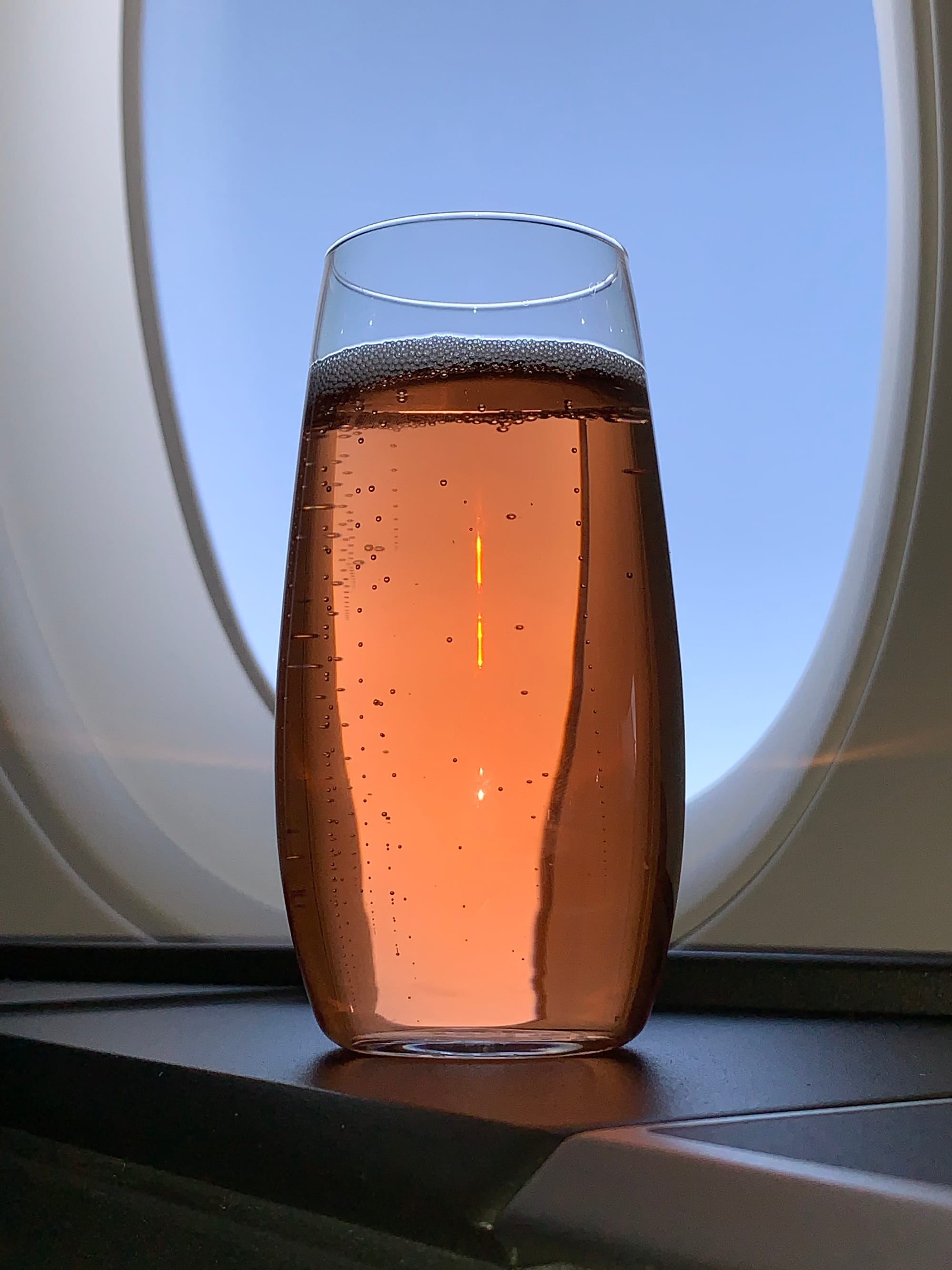 A stemless glass of pink bubbly in front of an airplane window.