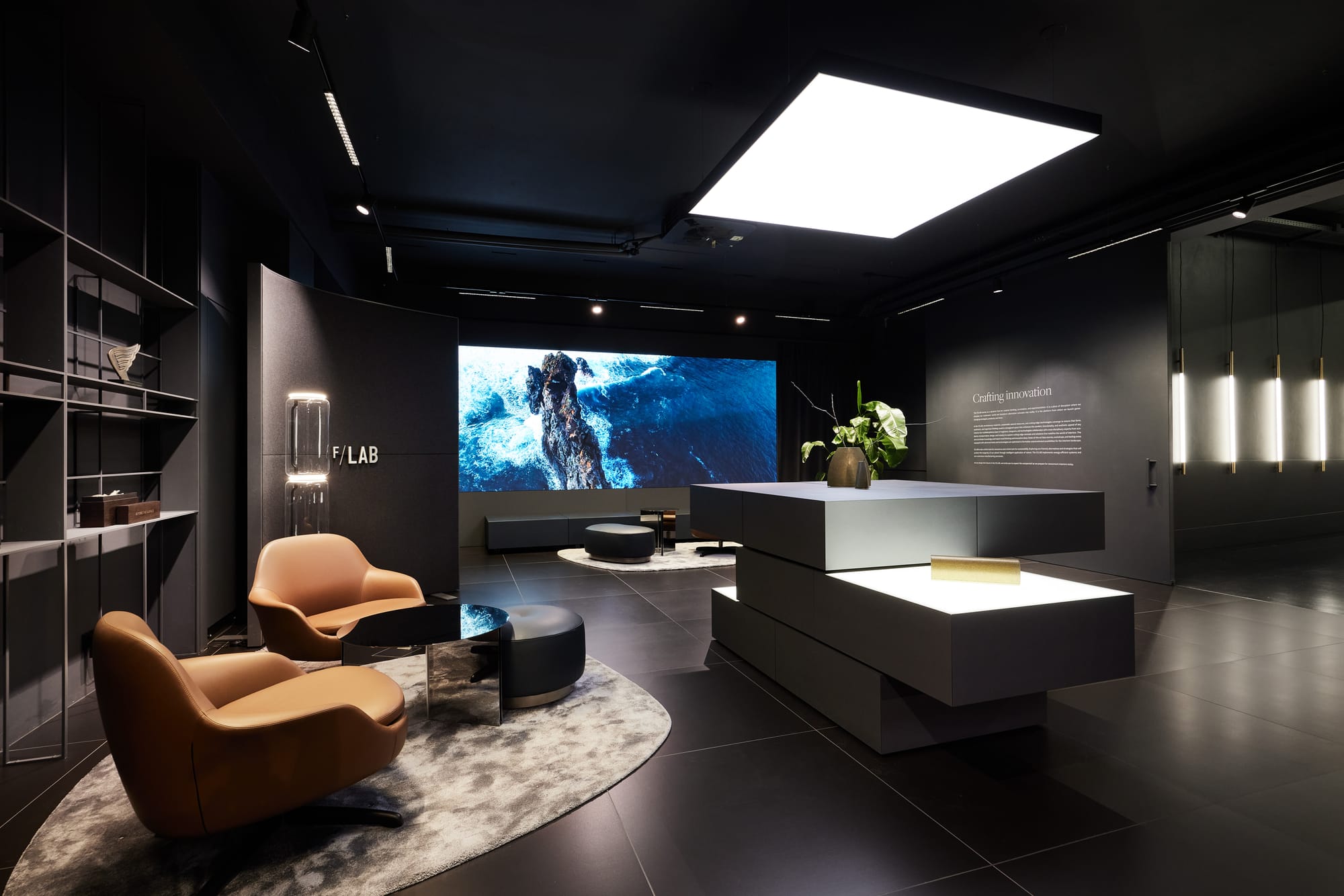 Interior space, mostly black walled modernist, with blocky elements and beige armchair seating. One wall is a video display. On another wall is marked "F/LAB".