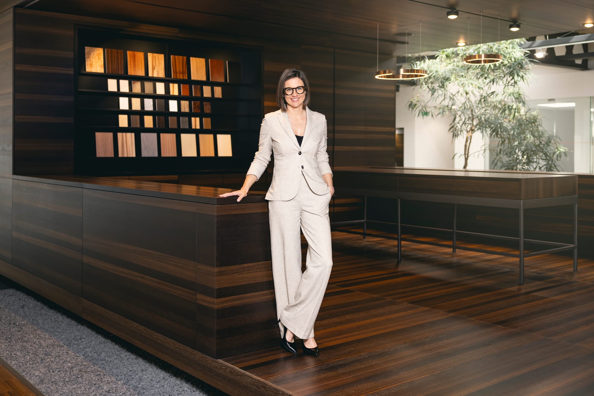 Interior, design lab. A woman in a tan suit stands on a wooden raised area, with various wood elements showcased behind her.